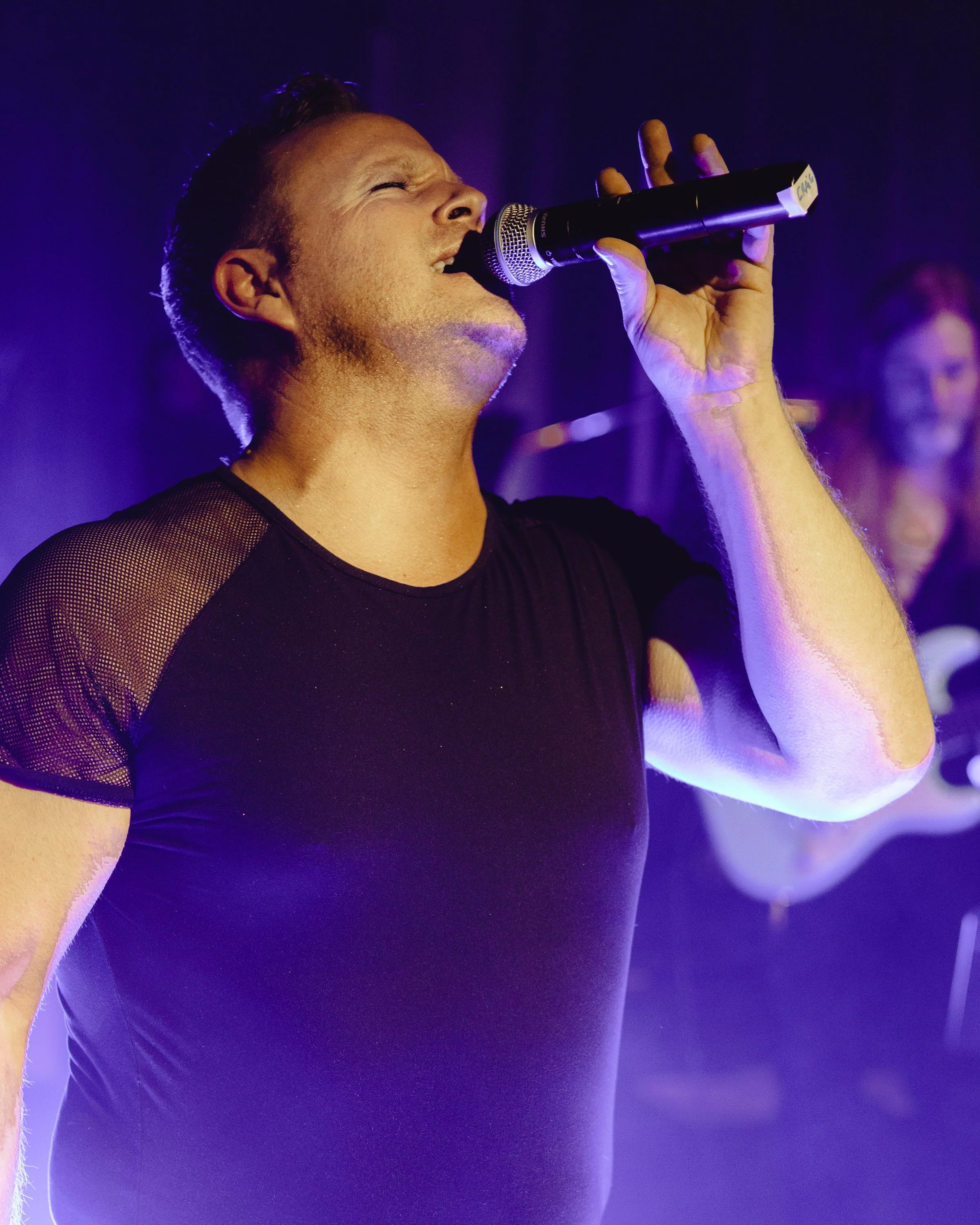 A person singing passionately into a microphone during a stage performance, wearing a black short-sleeved shirt. The setting is dimly lit with a blue-purple ambiance.