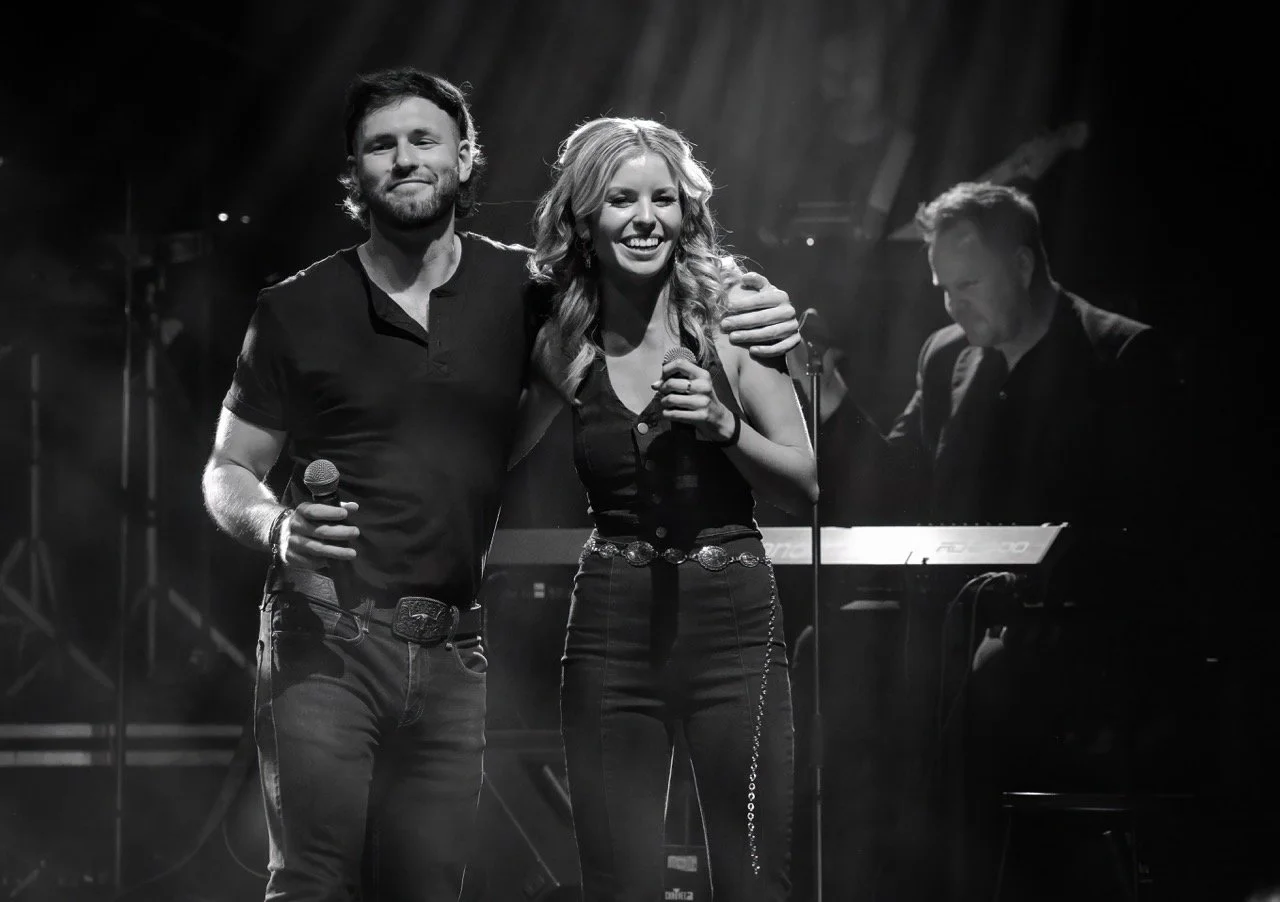 Black and white photo of two singers on stage, embracing with microphones; a keyboardist is visible in the background.