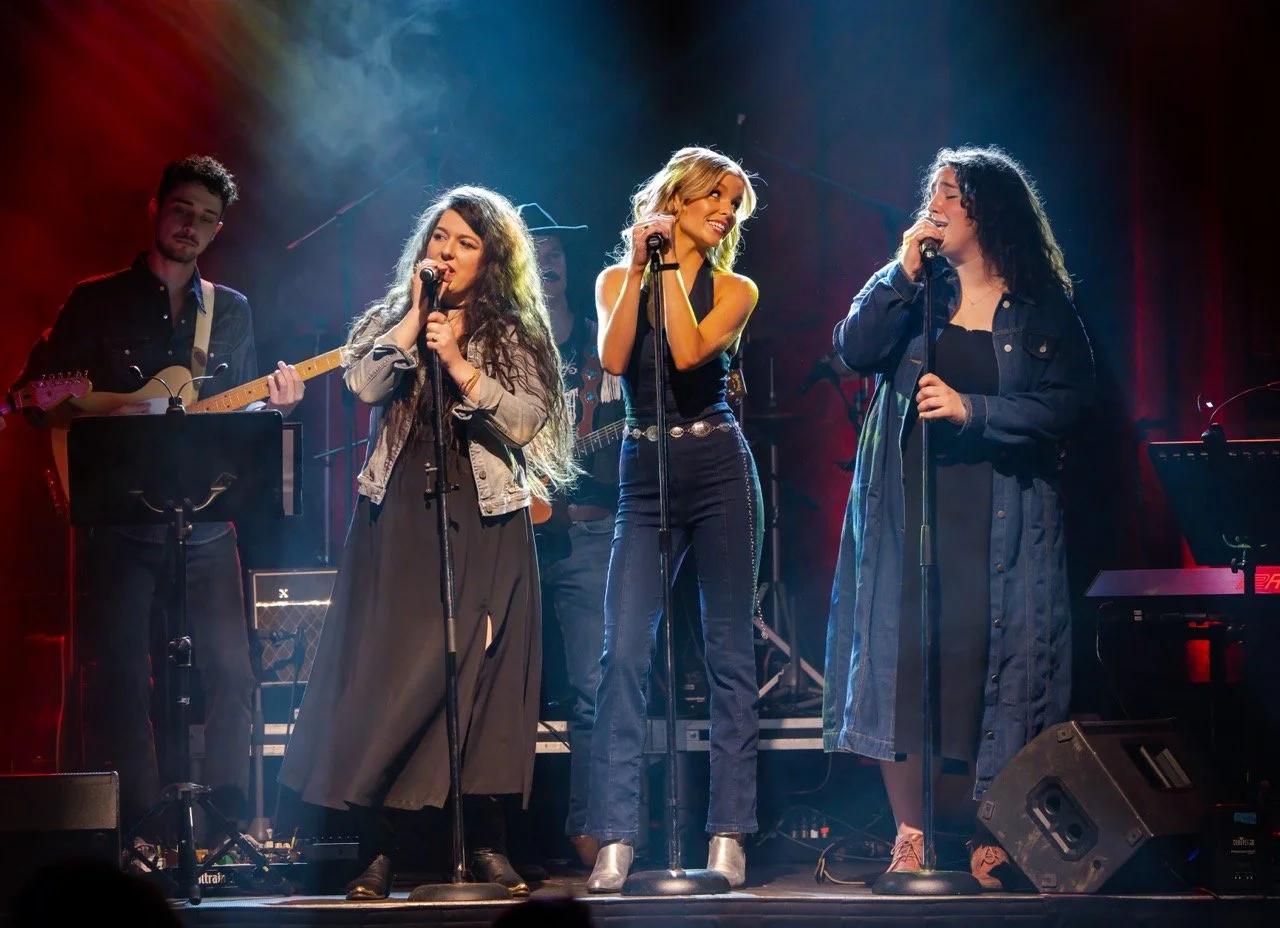 Group of musicians performing on stage, including three female singers and a male guitarist, under colorful stage lighting.