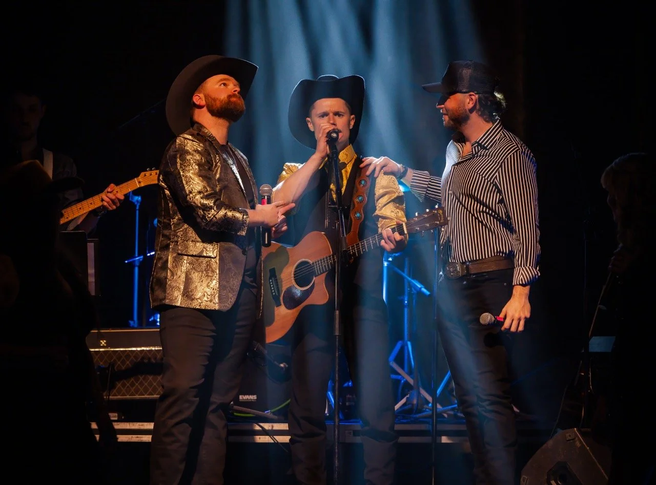 Three male musicians on stage, two holding microphones, one holding a guitar, wearing cowboy hats and country attire, blue stage lighting in the background.
