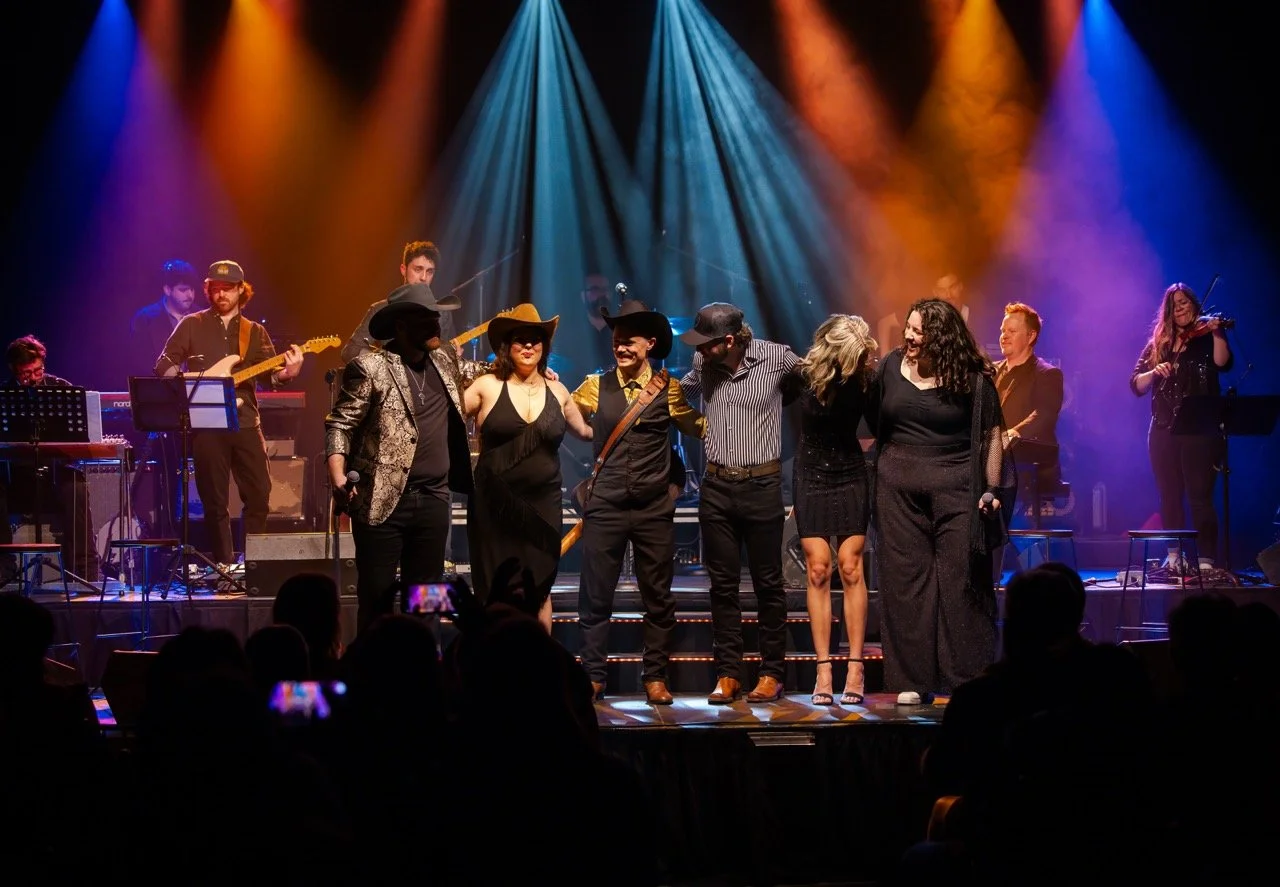 A group of people taking a bow on stage, with colorful stage lights and musicians playing instruments in the background.