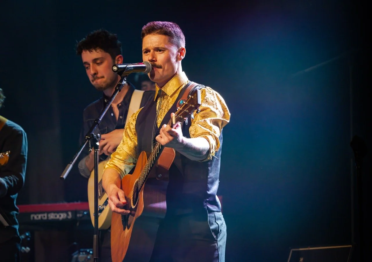 Musician performing on stage, playing guitar and singing into a microphone. Another person in the background playing an instrument, with stage lighting and keyboard visible.