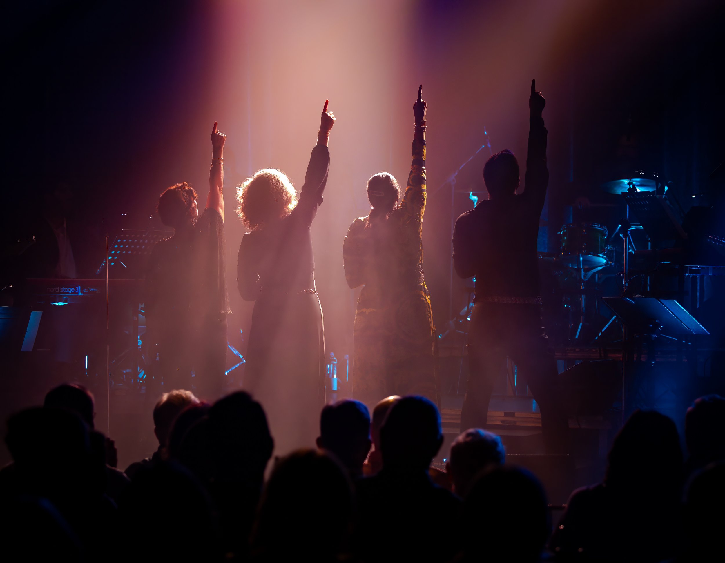 Four performers on stage facing a dimly lit audience, with colorful lighting and musical instruments in the background.