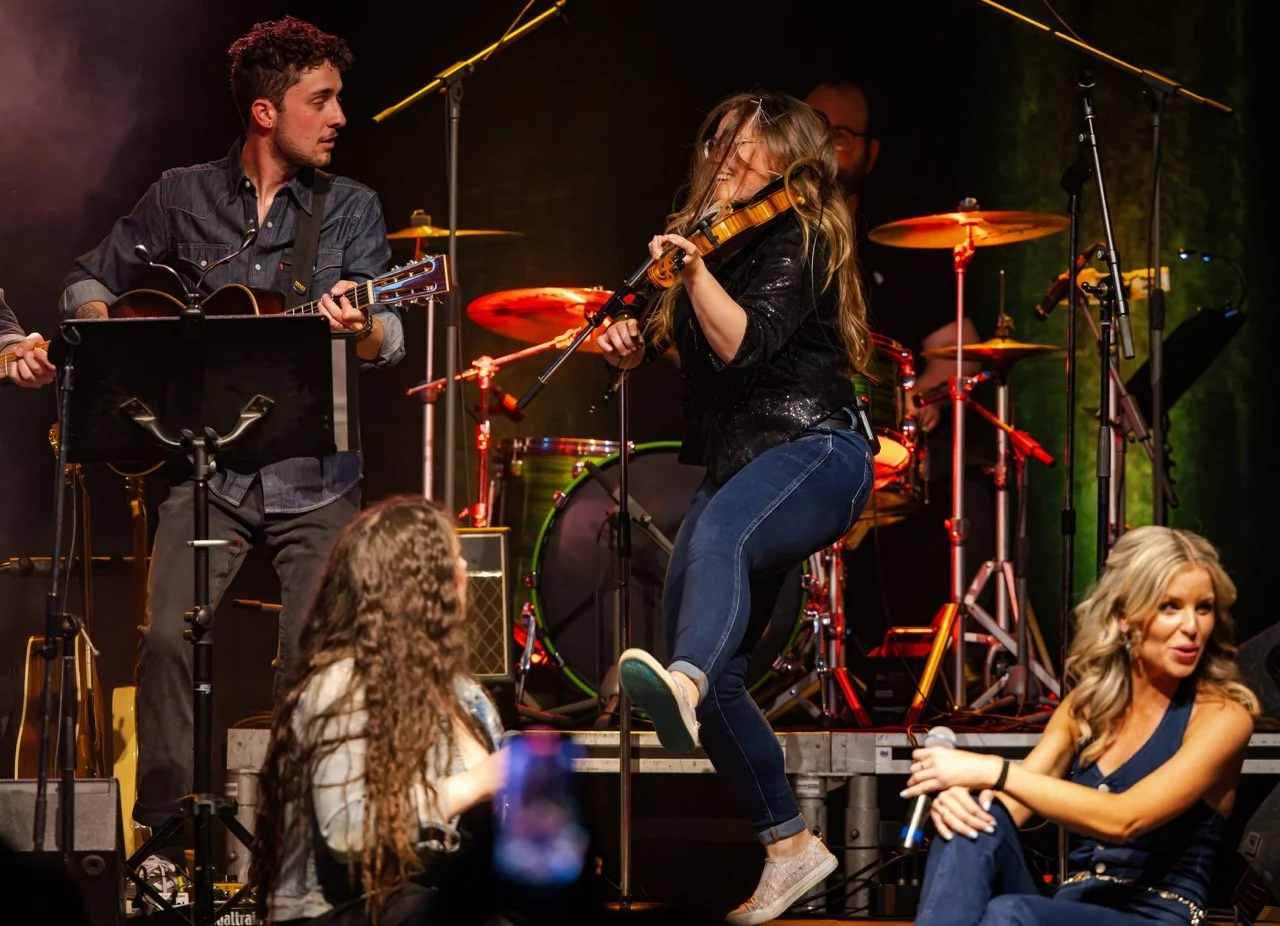 Band performing on stage with a guitarist and a violinist energetically playing. A female vocalist sits near the stage holding a microphone.