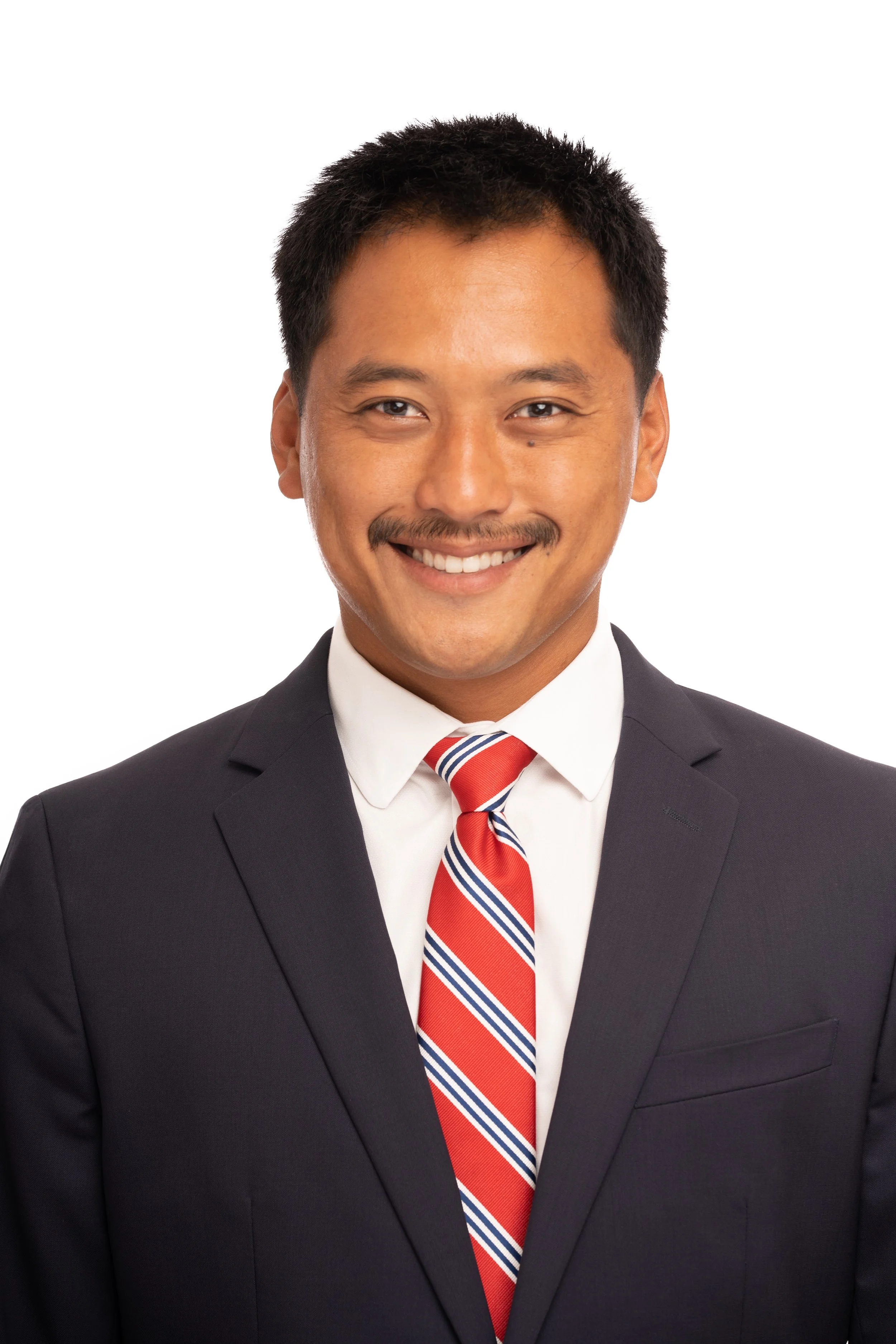 Professional headshot of a man with short dark hair and a mustache, wearing a dark suit jacket, white dress shirt, and red striped tie. He is smiling and looking directly at the camera against a plain white background