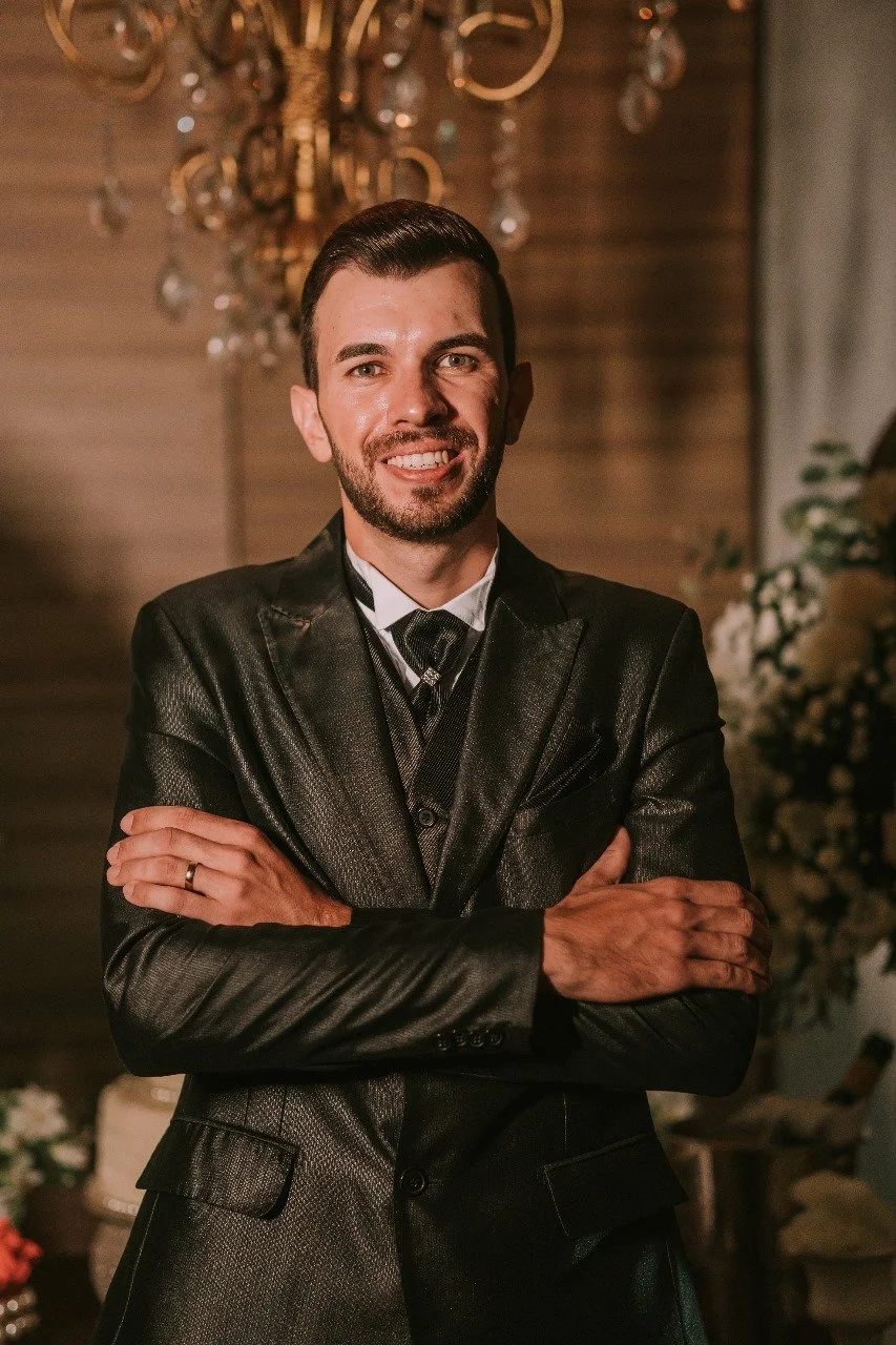 Homem sorridente com barba, cabelo penteado para trás, usando terno de couro preto, em ambiente com lustre e decoração floral ao fundo.