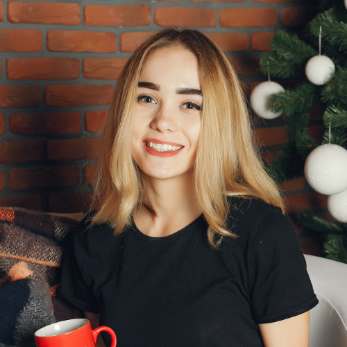Jovem sorridente com cabelo loiro falar na câmera, vestindo uma camiseta preta, ao lado de uma árvore de Natal decorada com bolas brancas, sentada em um ambiente com parede de tijolos vermelhos.