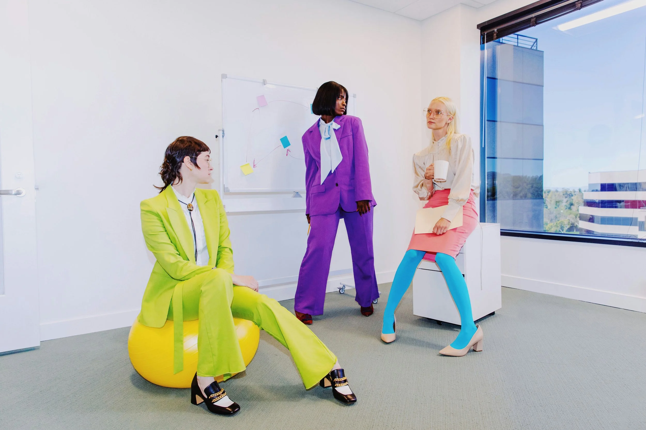 Four women in colorful, fashionable clothing in a modern office with large window, white walls, and whiteboard.