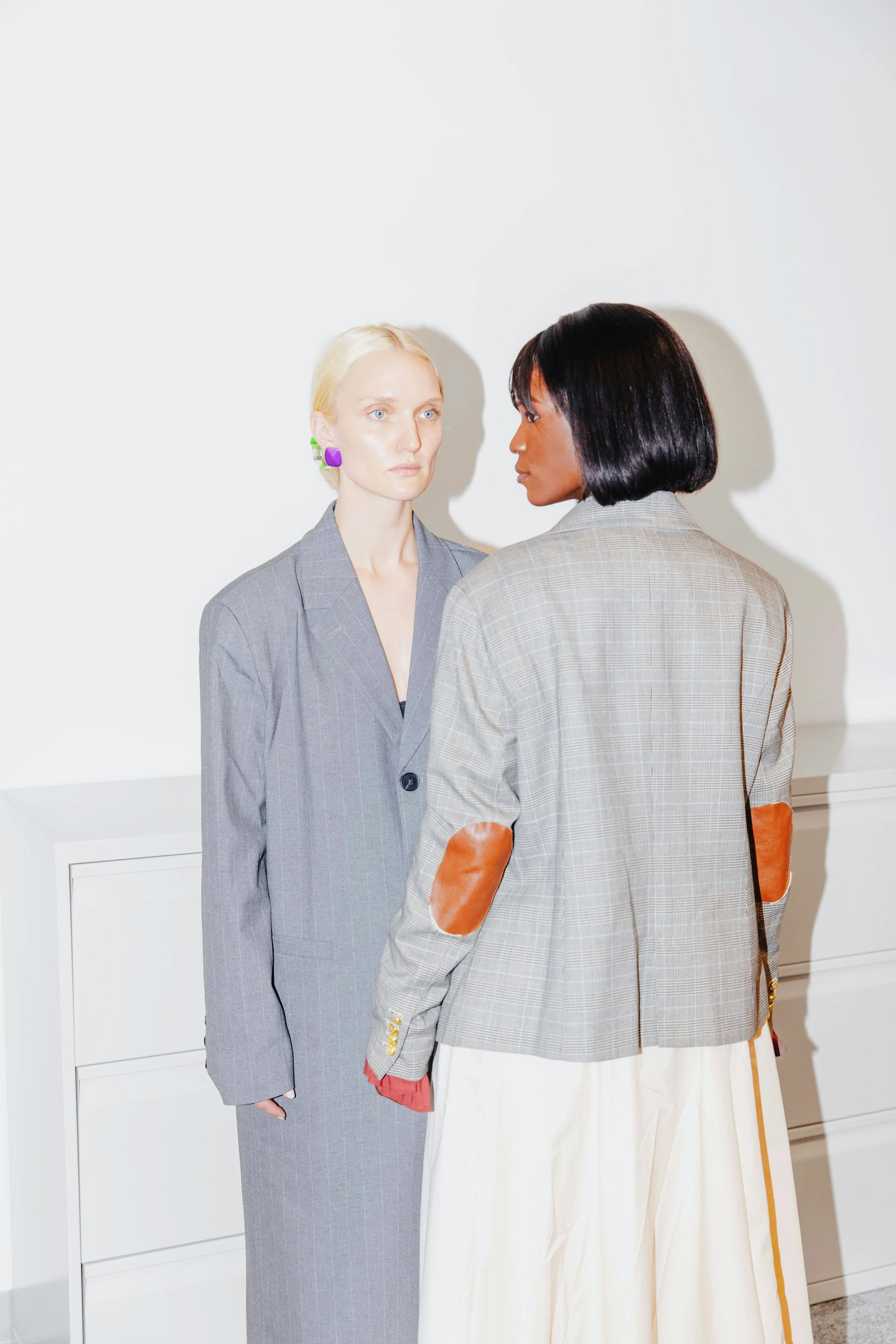 Two women in suits, facing each other closely, in a minimal white room. One woman has blonde hair and wears purple and green earrings, dressed in a gray pinstripe suit. The other woman has dark, shoulder-length hair and wears a beige plaid blazer with brown elbow patches, over a long white skirt.