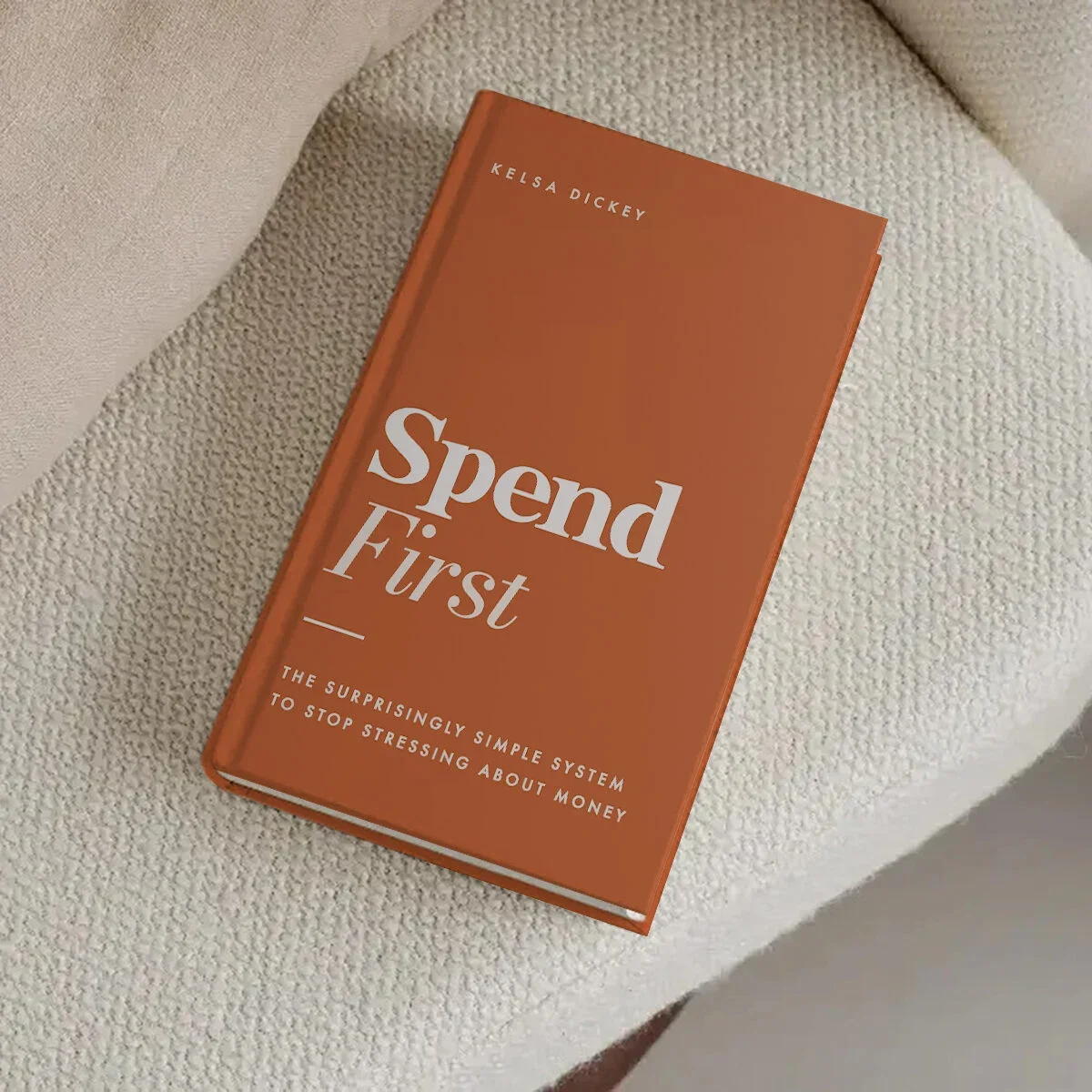 A book titled 'Spend First' by Kelsa Dickey resting on a cream-colored textured armchair.