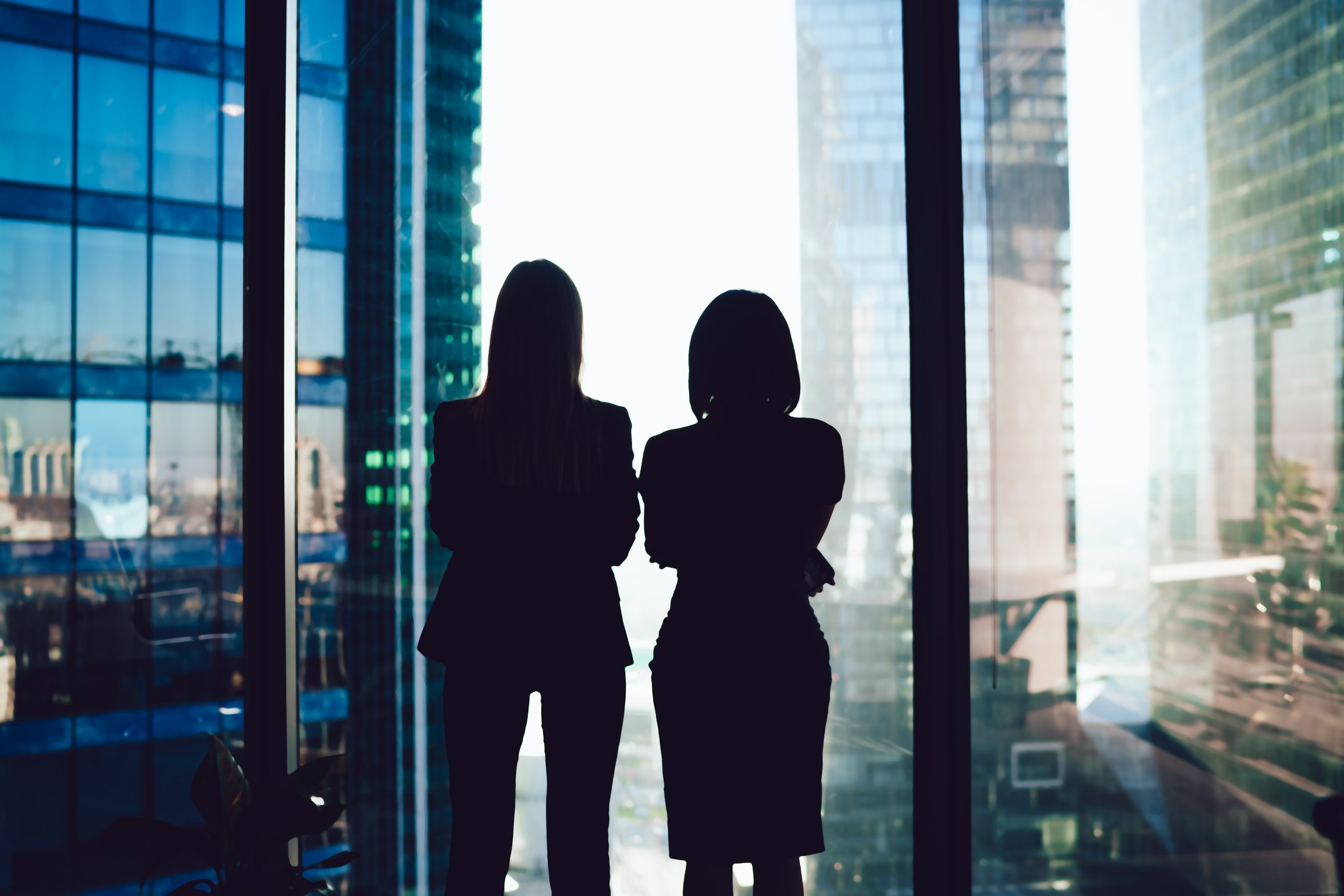 Two women in business attire standing in front of a large window overlooking a cityscape with high-rise buildings.