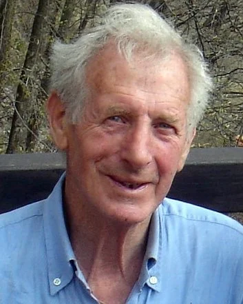 Uomo anziano con capelli grigi, sorriso, indossa camicia azzurra, scenario naturale sullo sfondo.