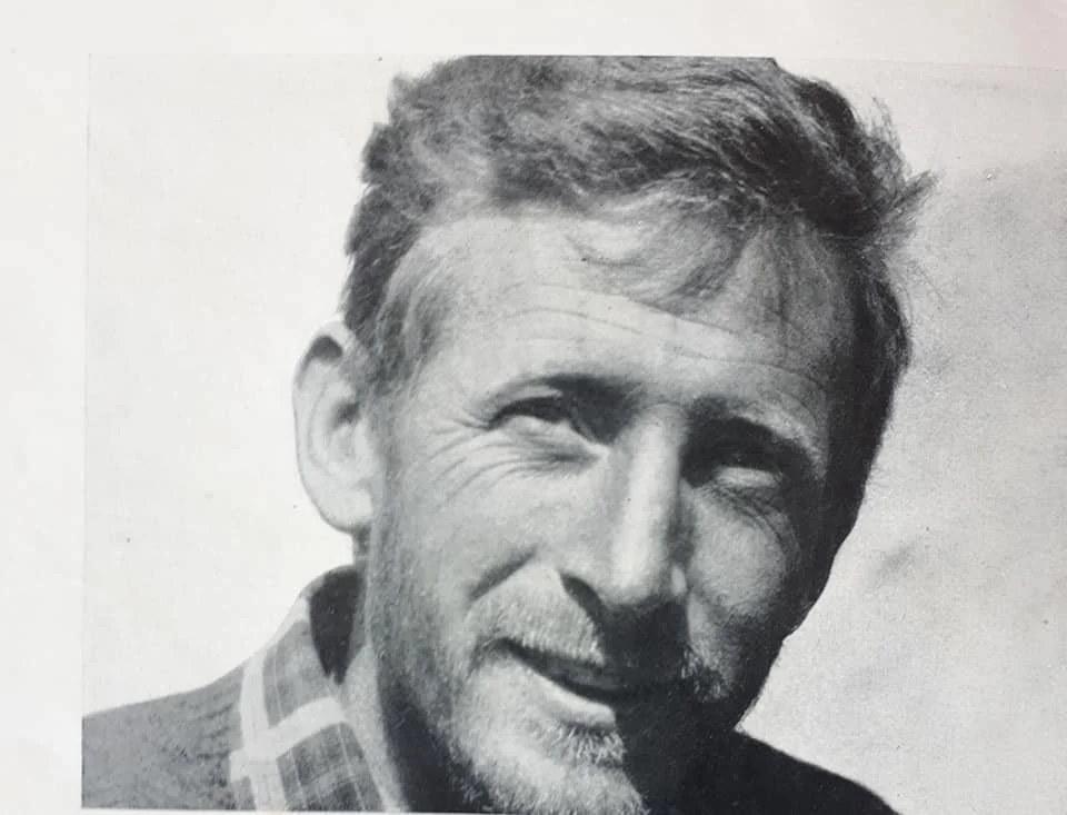 Ritratto in bianco e nero di un uomo con capelli castani, sorridente, con barba leggera, indossa una giacca con camicia a quadri.