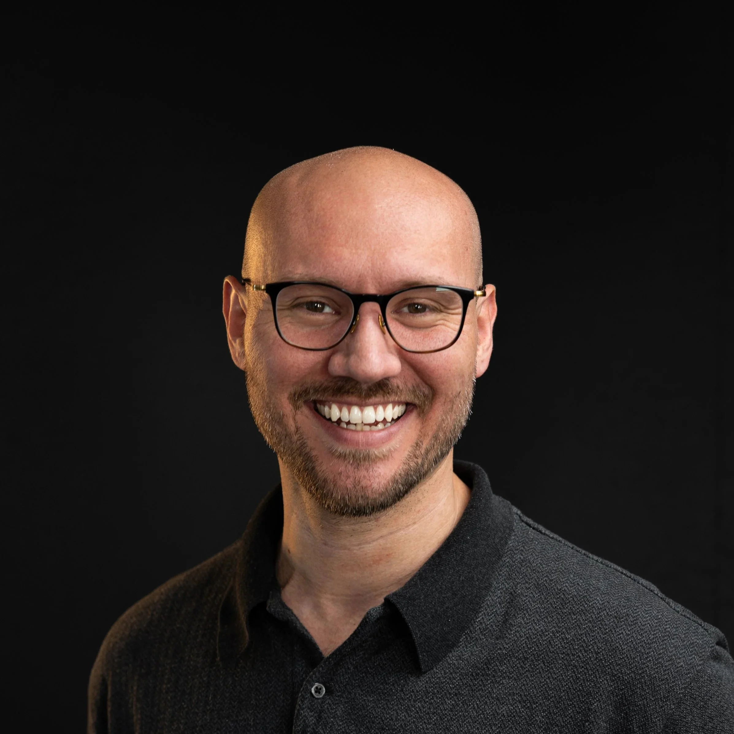Head of a smiling man with glasses and a beard, wearing a dark gray sweater, against a black background.