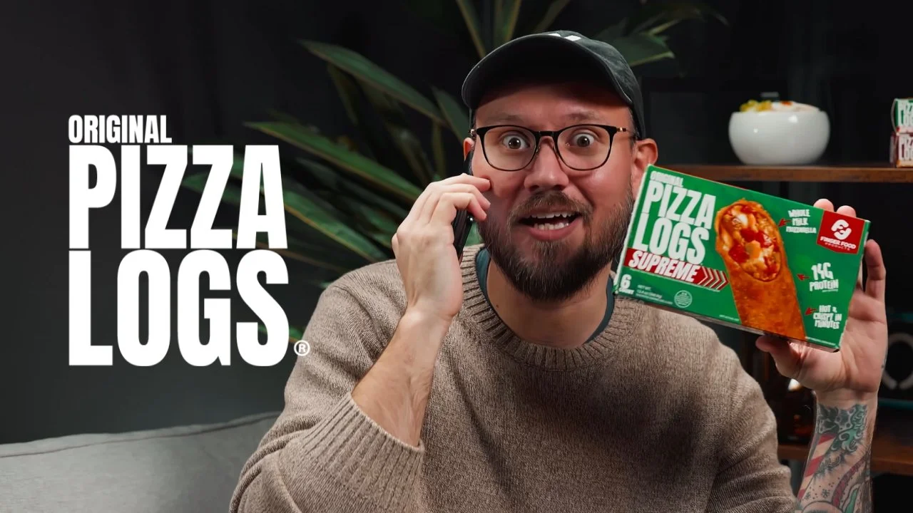 A man with glasses, a beard, and tattoos holding a box of Pizza Logs Supreme, talking on the phone, in front of a black background with large white text that reads 'Original Pizza Logs'.