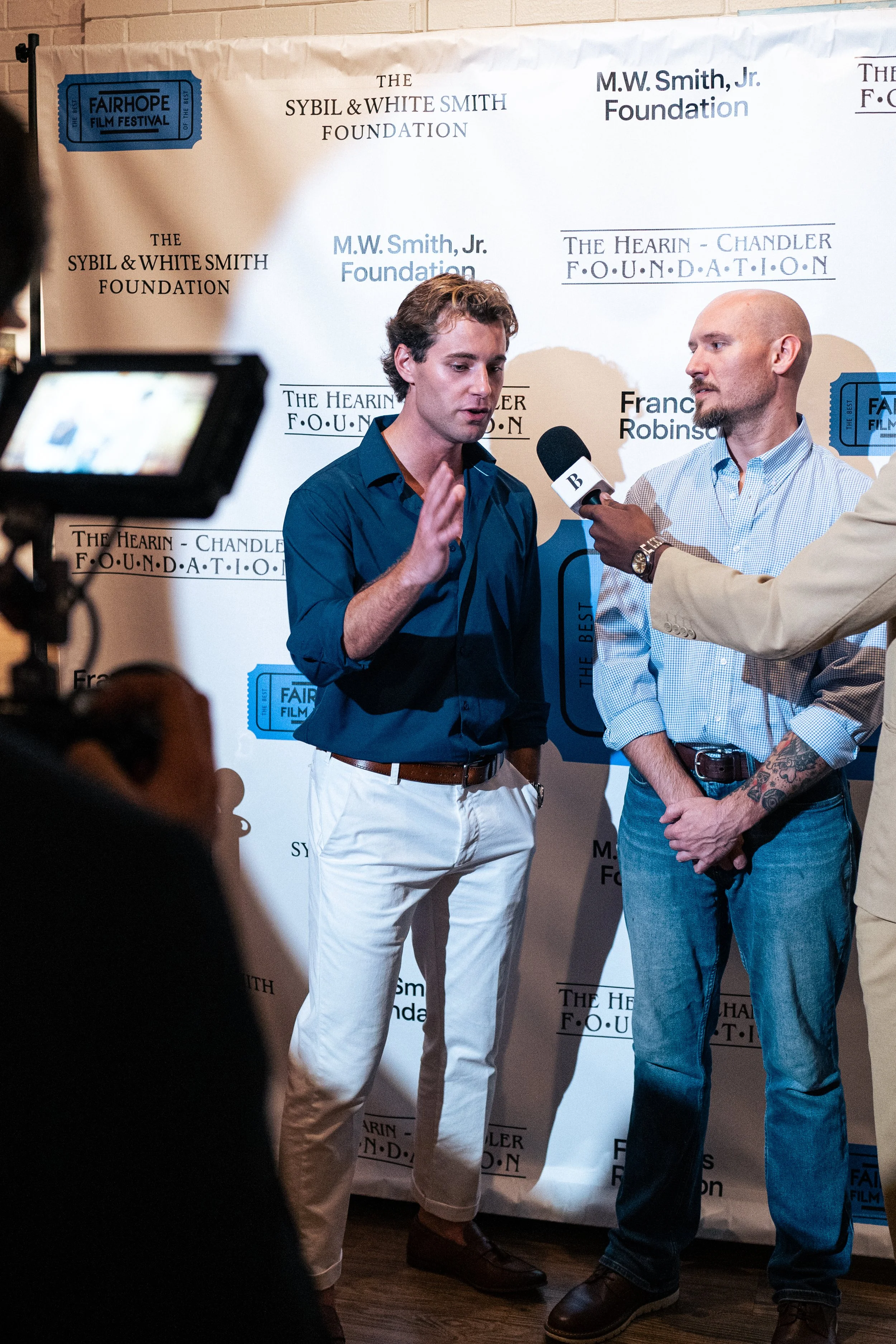 Two men are having an interview on a red carpet at an event with a backdrop displaying multiple logos and text, including 'The Sybil & White Smith Foundation,' 'M.W. Smith Jr. Foundation,' and 'Fairhope Film Festival.' One man is speaking into a microphone held by a third person, and the other man is listening attentively.