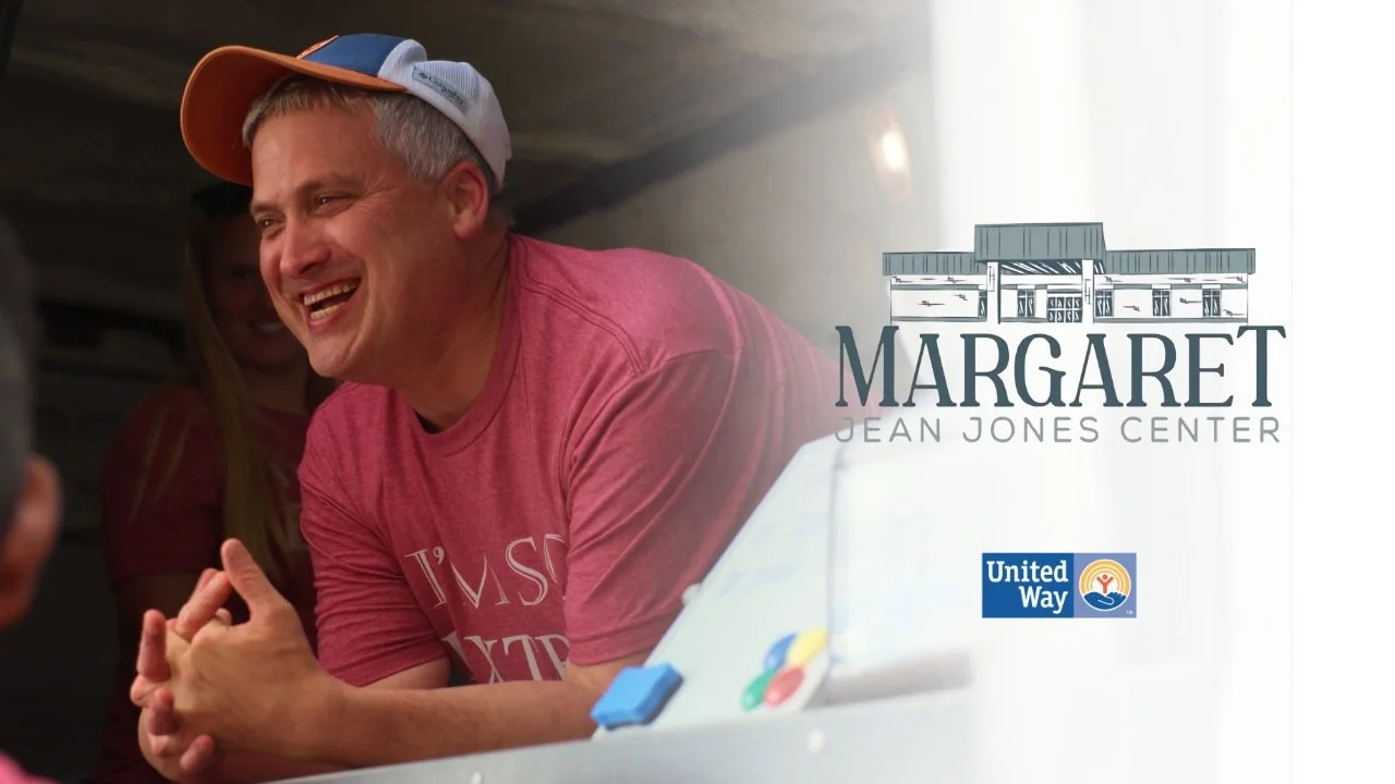 A man wearing a colorful cap and pink t-shirt smiling and engaging in a conversation indoors, with a young woman in the background. The image includes the Margaret Jean Jones Center logo and United Way logo.