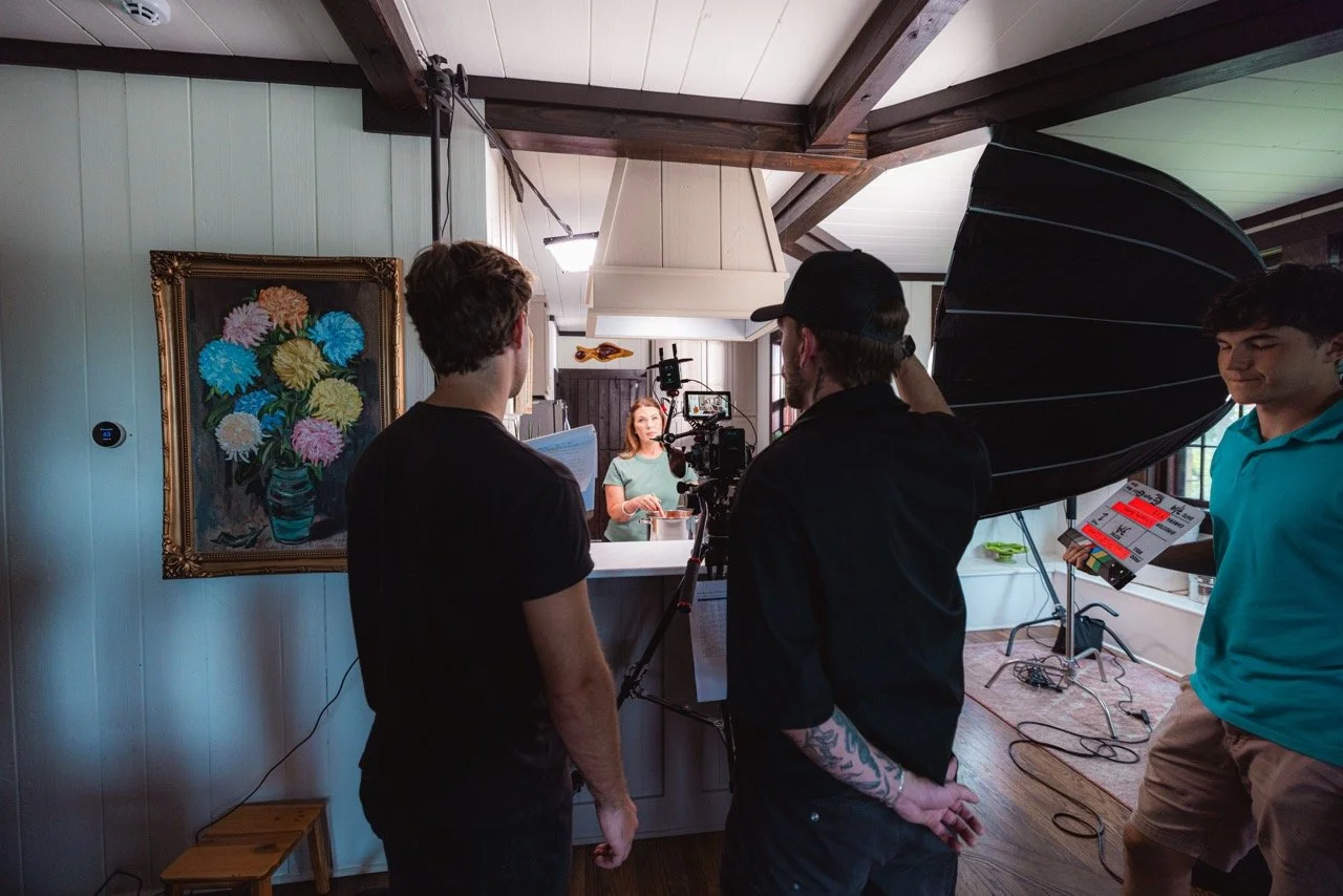 Filming crew working on a video shoot with a woman speaking in a kitchen. Crew members operate a camera and lighting equipment, with a clapperboard visible.