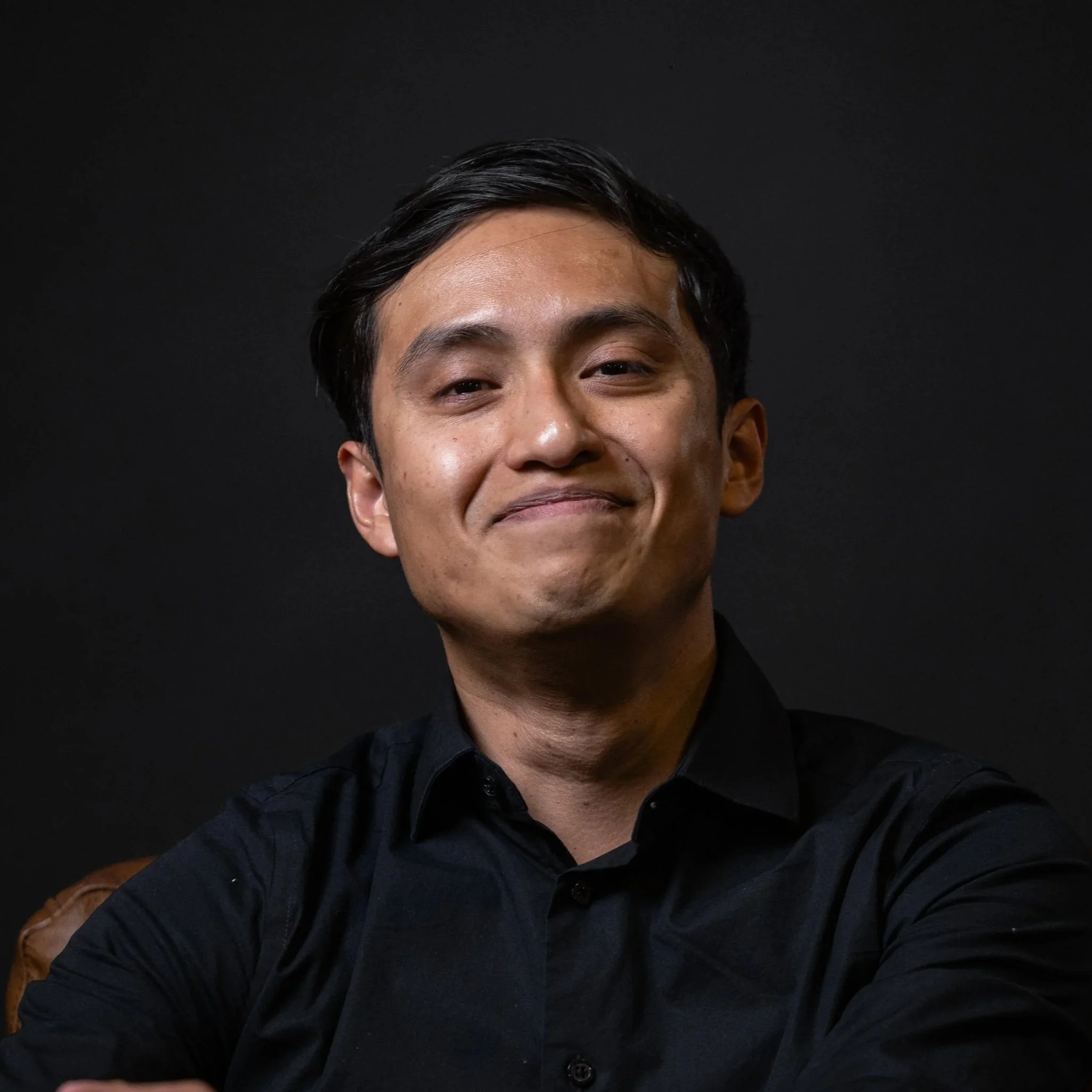 Close-up of a smiling man with dark hair in front of a black background.