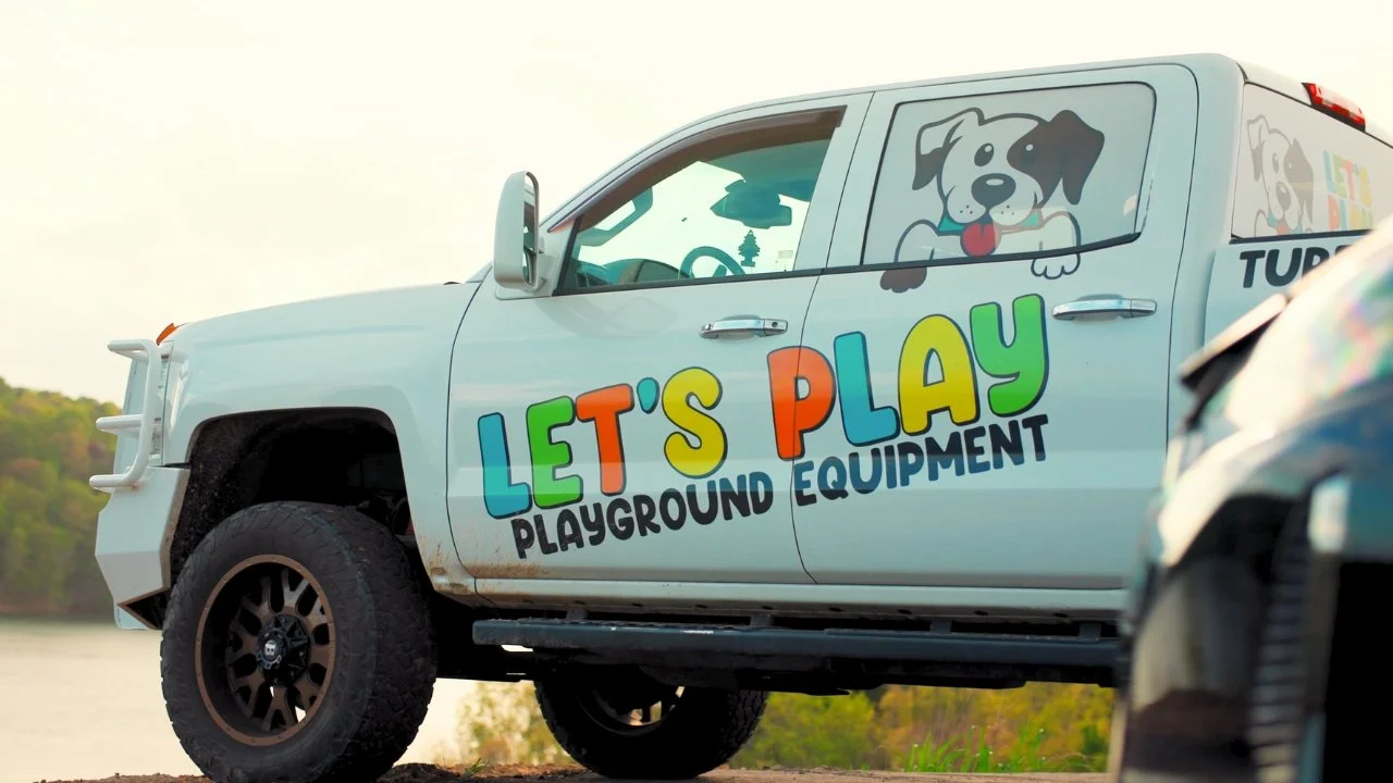 White pickup truck with colorful lettering and cartoon puppy on the side, advertising playground equipment for children, parked outdoors near a body of water and trees.