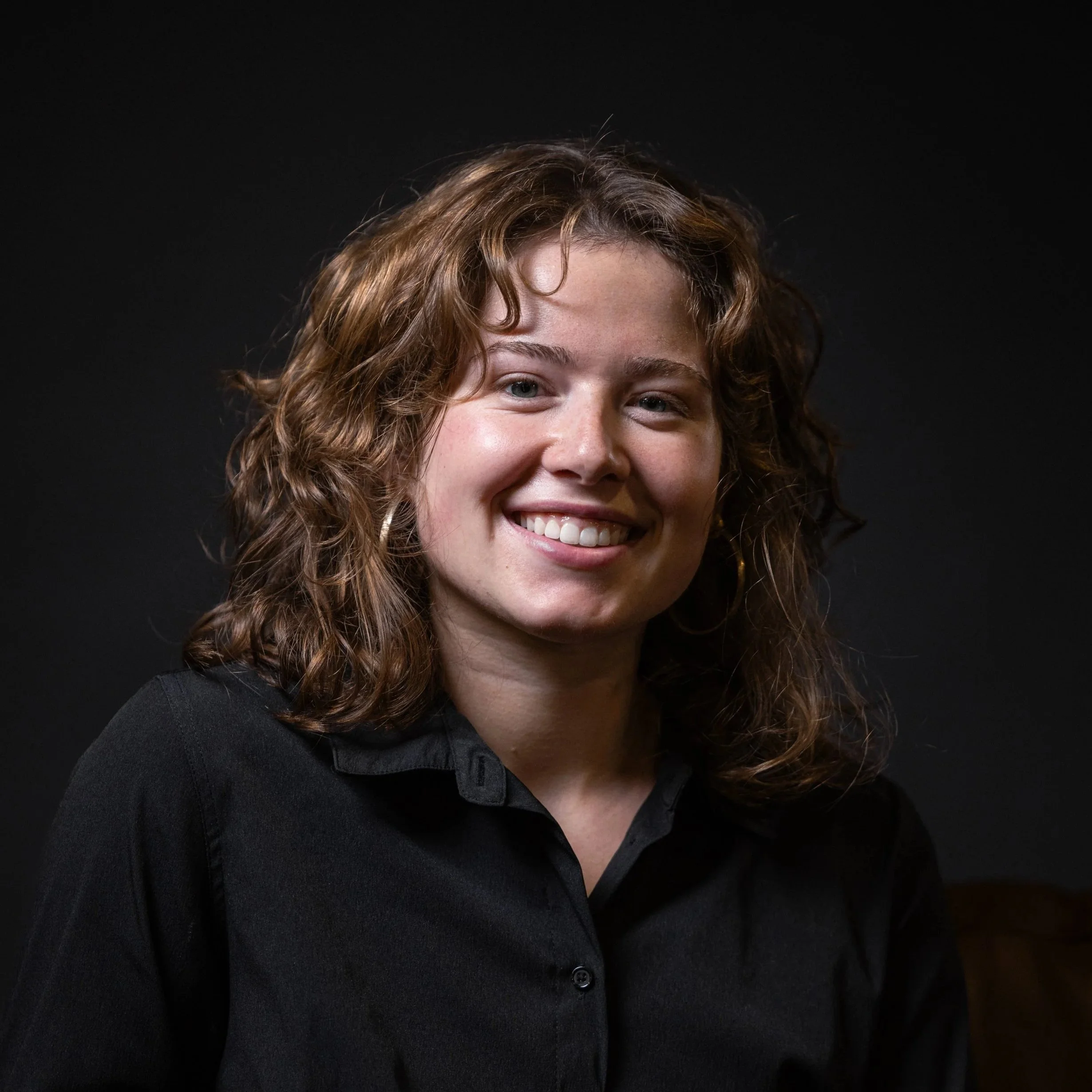 Close-up of a smiling woman with curly, light brown hair wearing a black shirt against a black background.