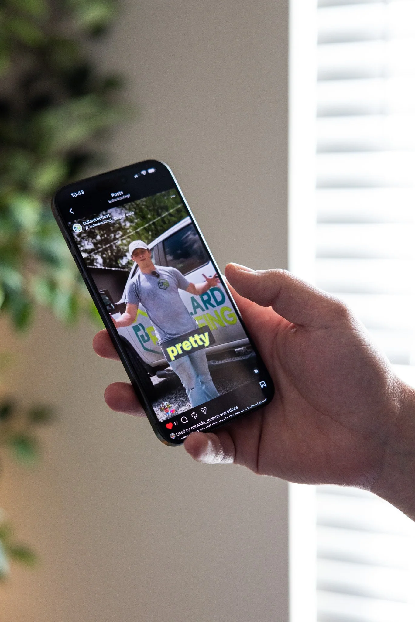 Close-up of a person's hand holding a smartphone displaying a social media post with a man standing outdoors near a vehicle, wearing a gray t-shirt and a white cap, with colorful text on the post.
