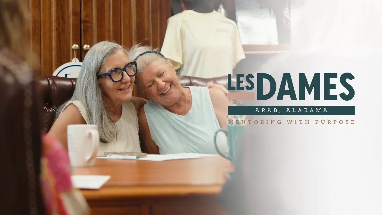 Two older women sharing a joyful moment, sitting at a table in a cozy room with wooden-paneled walls; one woman has long gray hair with glasses, and the other has short gray hair and a headband. Text overlay reads 'Les Dames Arab, Alabama Mentoring with Purpose'.