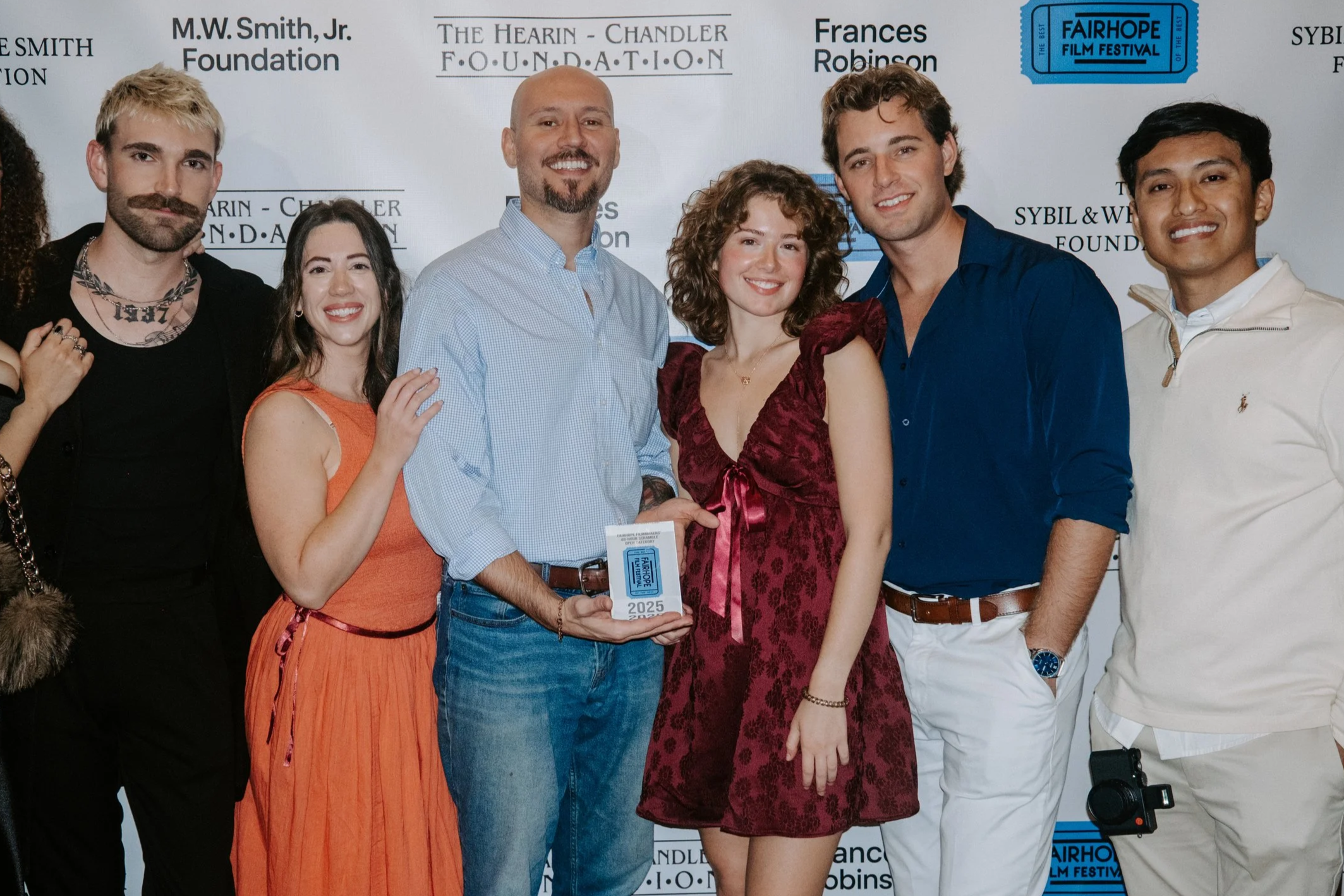 A group of six people standing together at a film festival event, with some holding an award, in front of a backdrop displaying various sponsors and event logos.