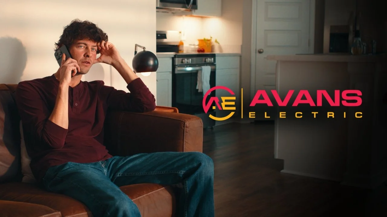 A man sitting on a brown couch, talking on a phone in a kitchen with a modern stove and some plants, with the