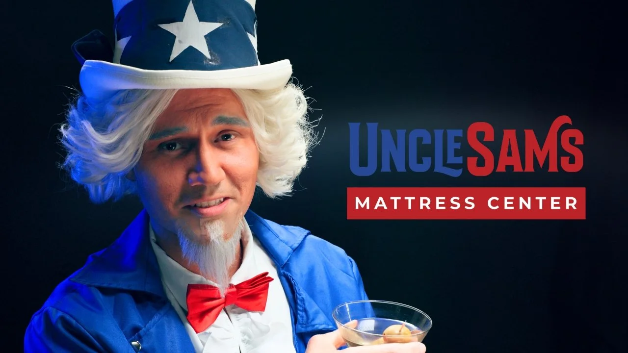 A man dressed as Uncle Sam, with a white wig, red bow tie, and a blue jacket, holding a glass of cocktail, standing next to a sign that says "Uncle Sam's Mattress Center."