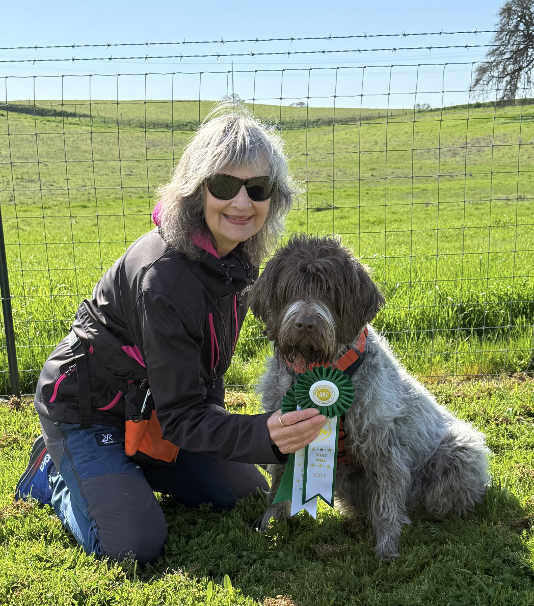 A person smiling, kneeling beside a dog while holding award ribbons. The dog has a shaggy coat and is sitting. They are outdoors, in front of a wooden wall.