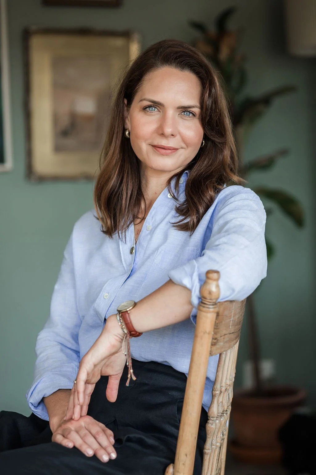 A woman with shoulder-length brown hair, blue eyes, and fair skin, sitting on a wooden chair with a relaxed posture, wearing a light blue button-up shirt and dark pants, with a background of framed artwork and green plants.