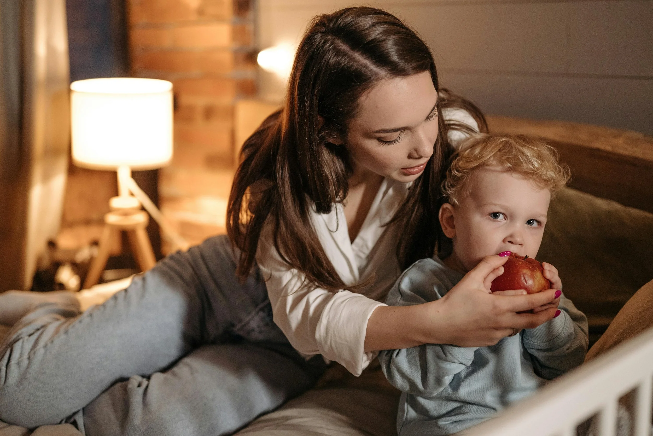 Mother sitting with young child during calm bedtime routine as child eats apple in peaceful evening setting, showing predictable routines that help managing kids emotions during celebrations by maintaining stability