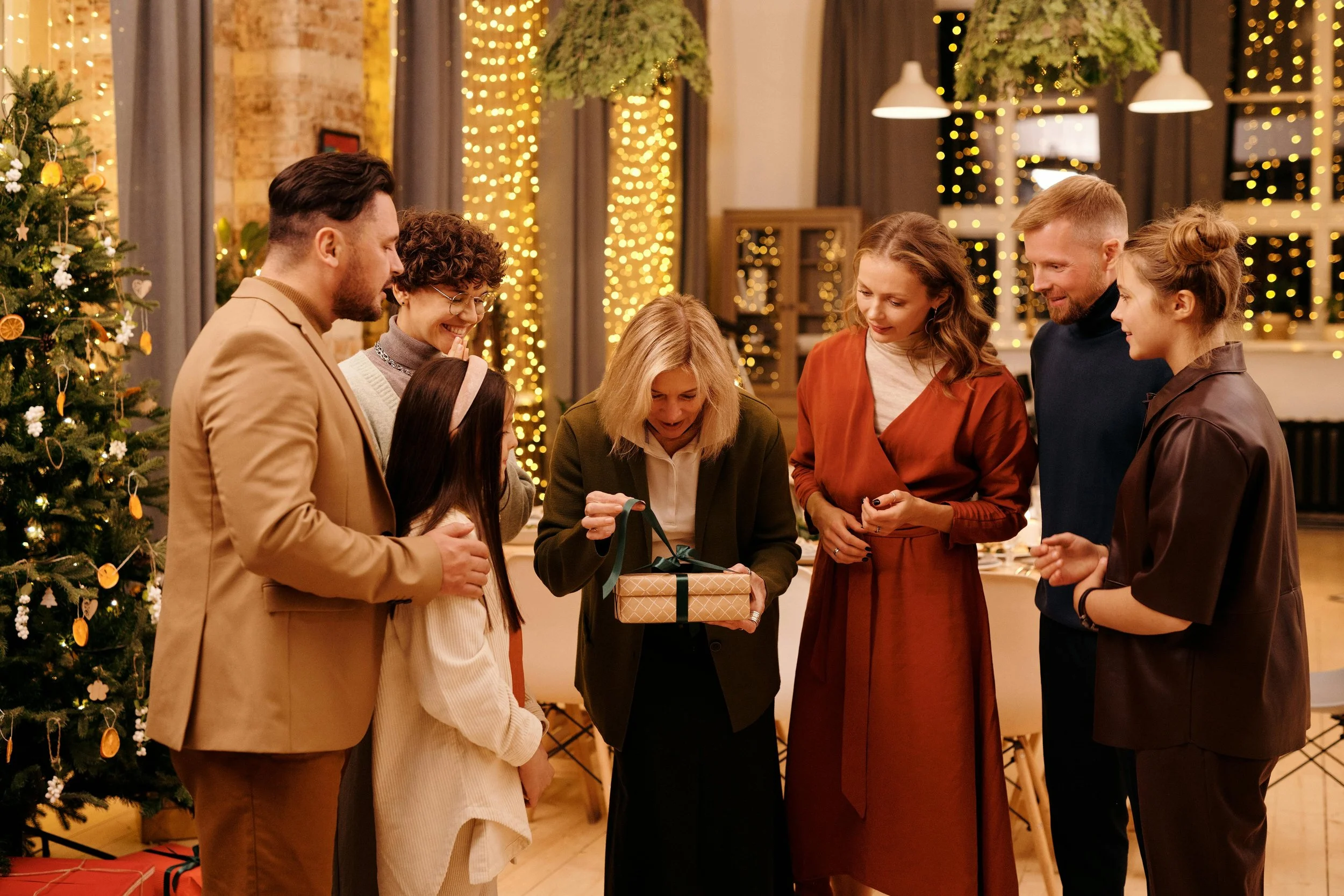 Adults and a kid gathered at festive holiday party with Christmas trees watching gift exchange, illustrating how parents can model calm New Year anxious kids support by managing their own holiday expectations and transitions