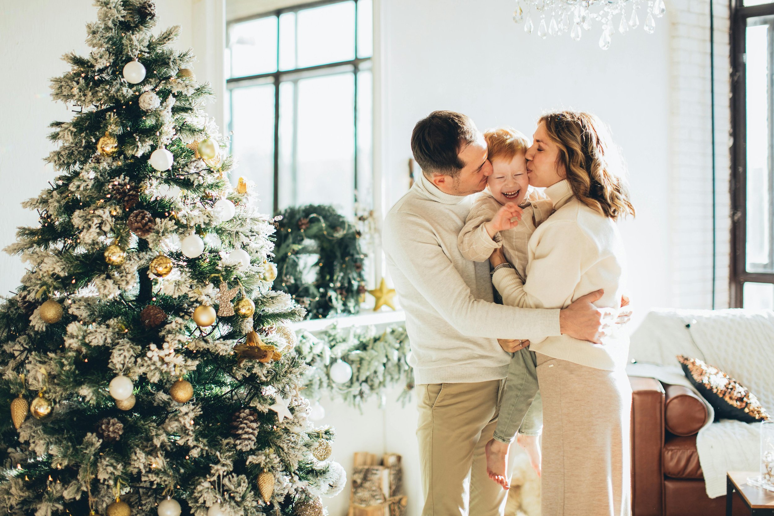 Parents kissing laughing child on both cheeks beside decorated Christmas tree, capturing how reducing holiday stress children happens through genuine affection, connection, and joyful family traditions