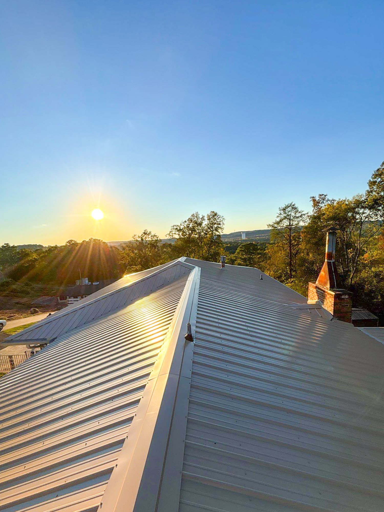 View of a house roof with metal panels during sunset, with trees in the background and a chimney on the right side.