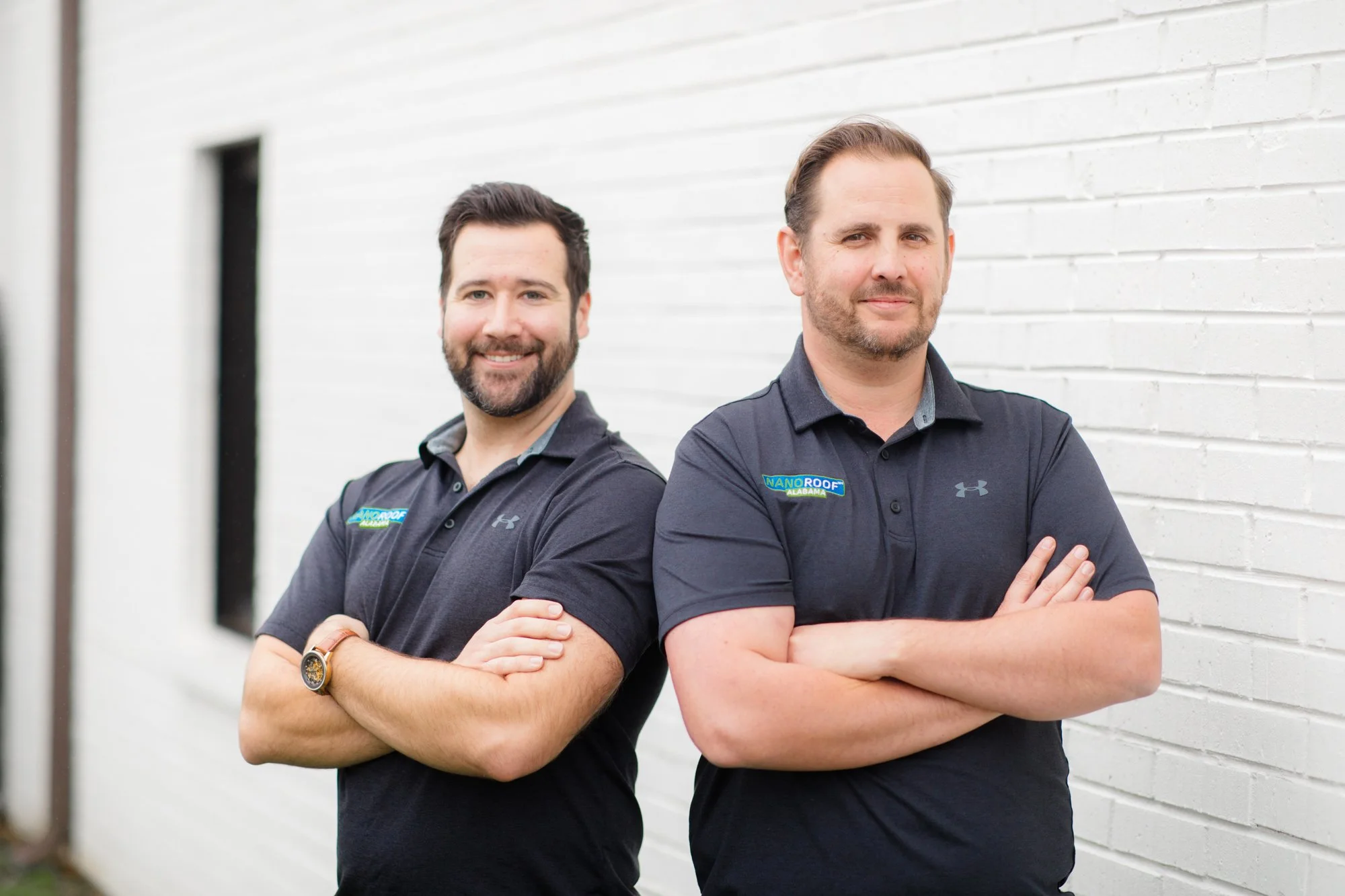 two men, the owners of NANOROOF Alabama, smiling with their arms crossed wearing NANOROOF Alabama branded grey polos and standing in front of a white brick wall