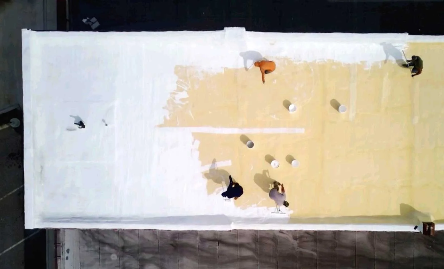 Aerial view of a spray foam coated flat roof being coated with high solids silicone sealant by men with rollers and buckets.