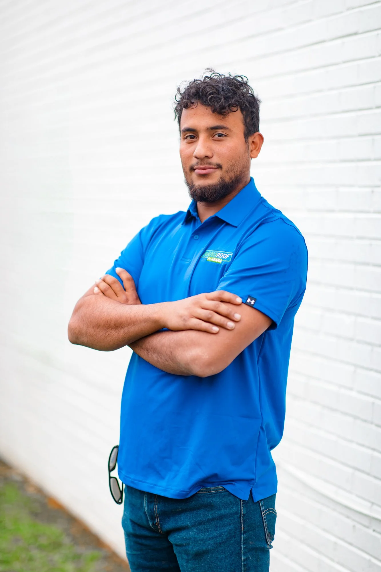 A man wearing a blue polo with a NANOROOF Alabama logo, with his arms crossed, slightly smiling, against a white brick wall background 