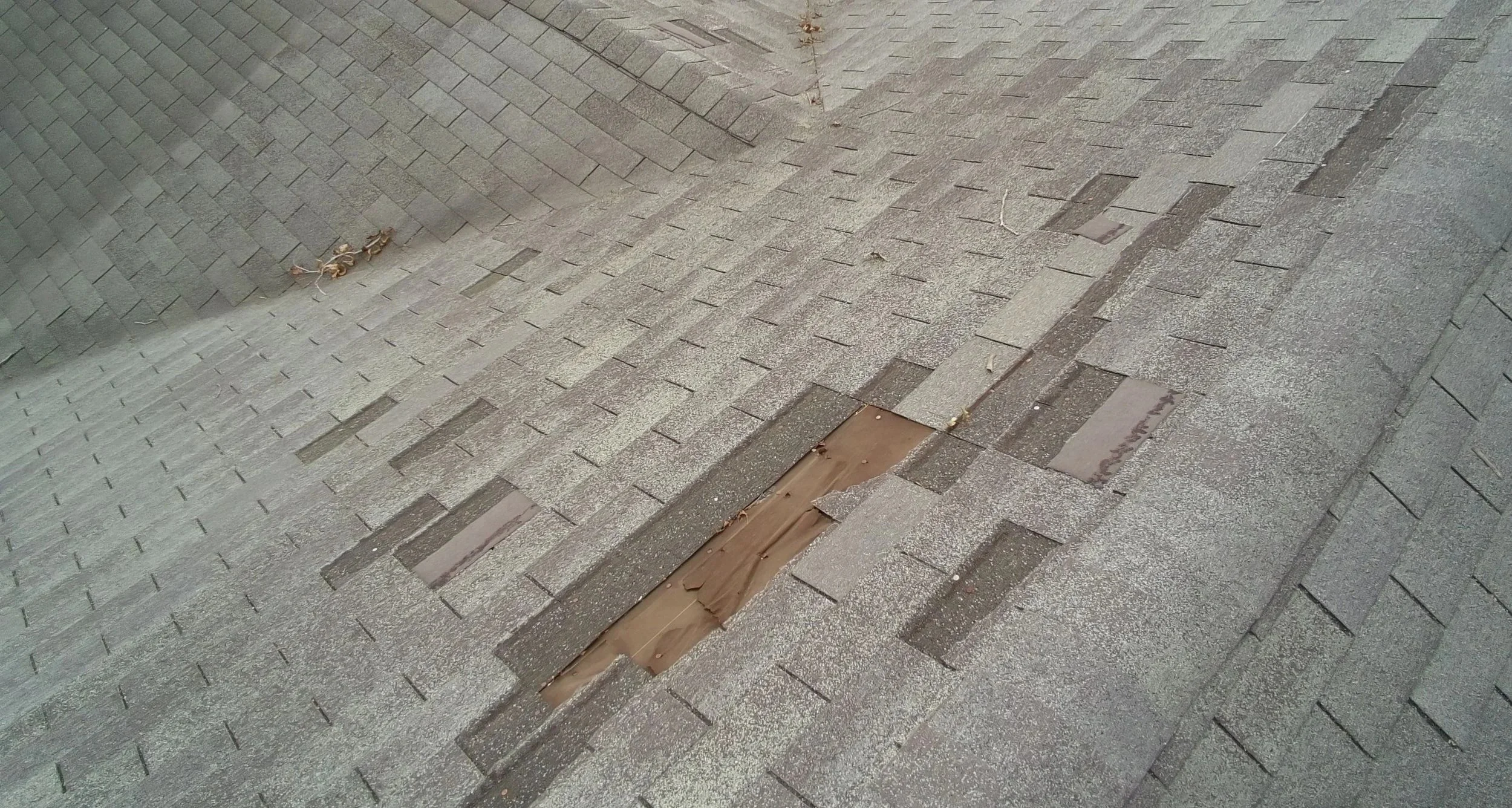 view of top of an asphalt roof with damaged and missing shingles, exposing the brown underlayment