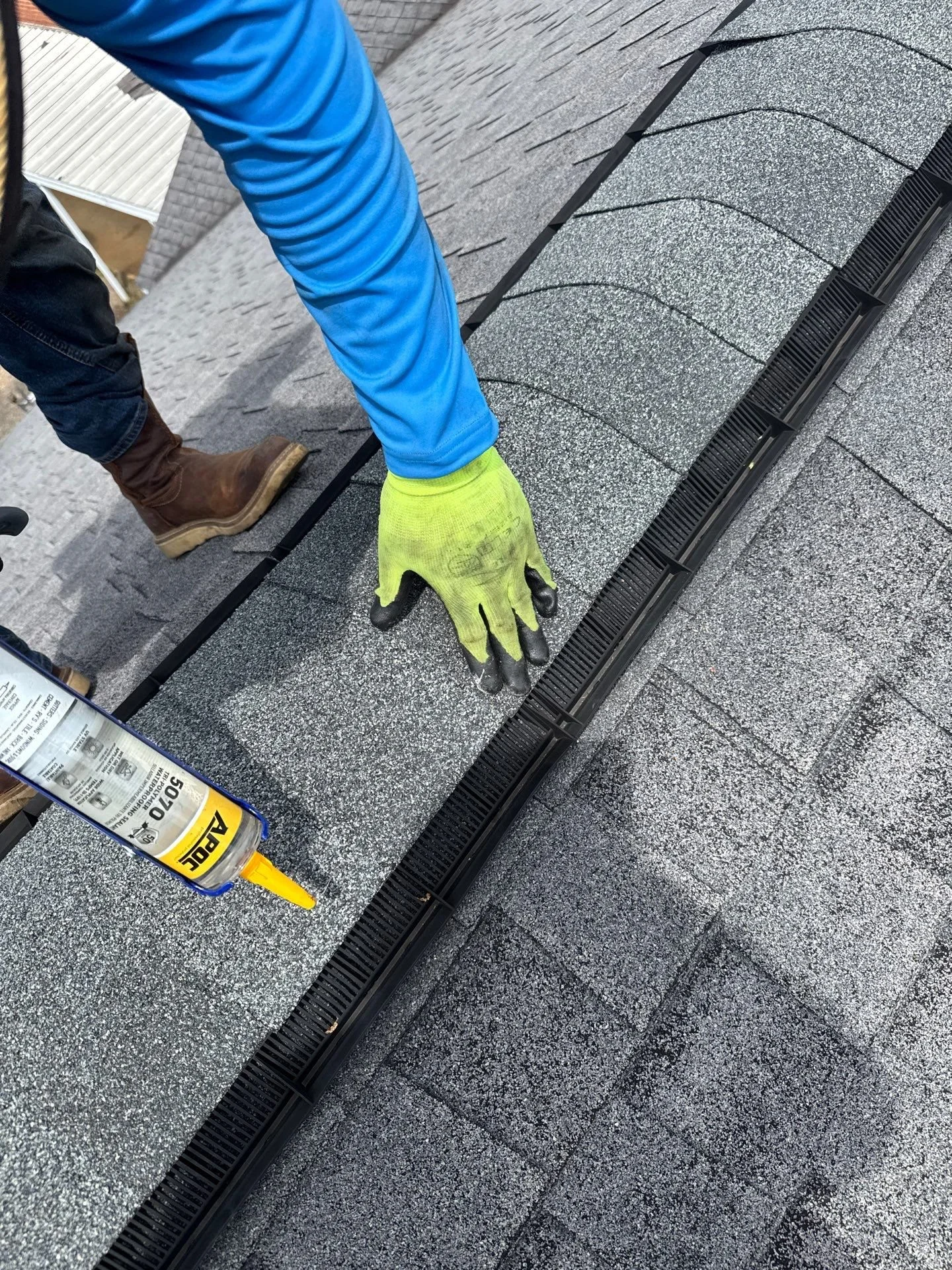 Aerial close up view of a roofer applying caulk to a ridge vent, wearing a blue long sleeved shirt with a neon green and black glove