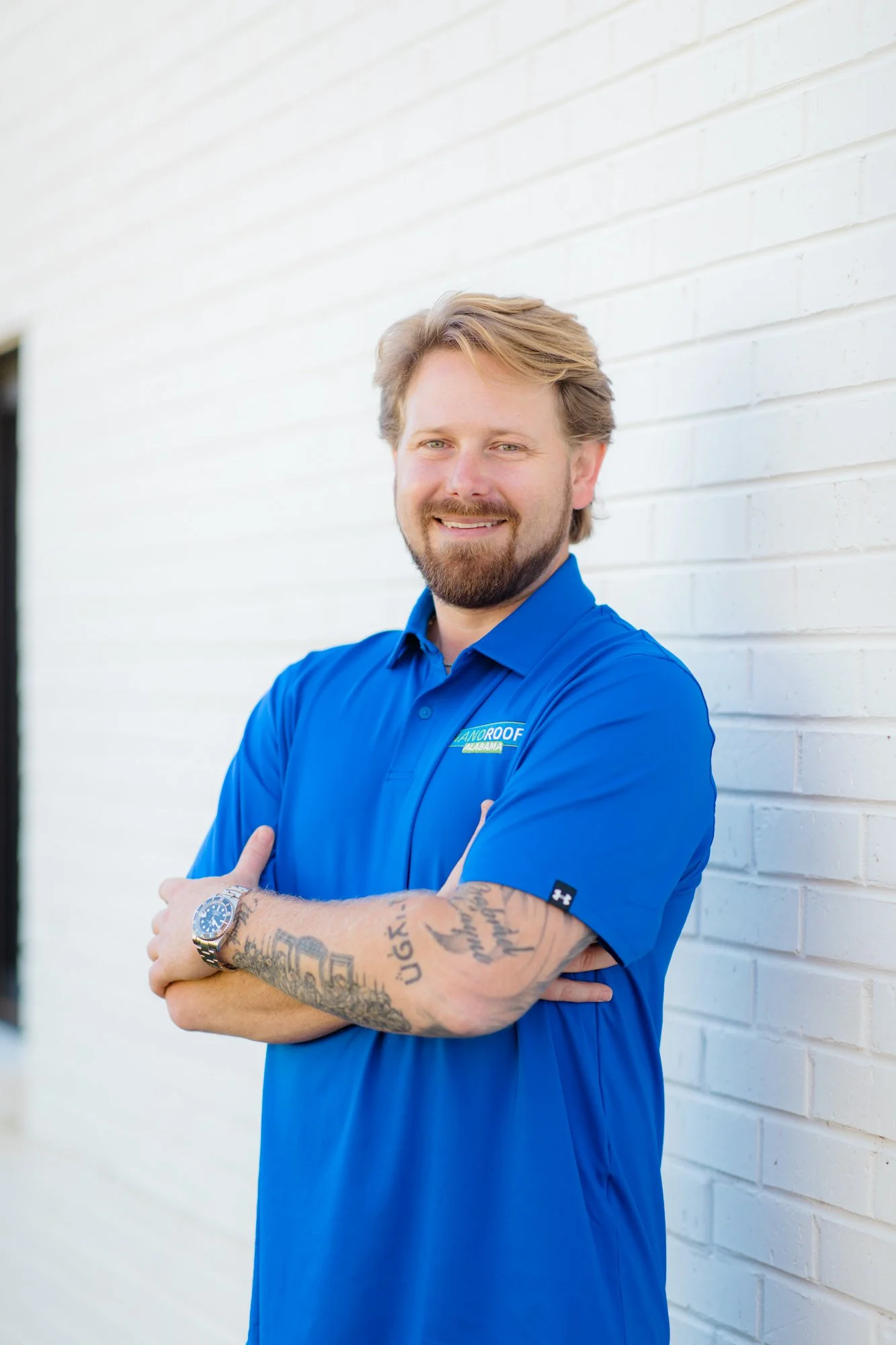 A smiling man with red hair wearing a blue polo with a NANOROOF Alabama logo, with his arms crossed against a white brick wall background 