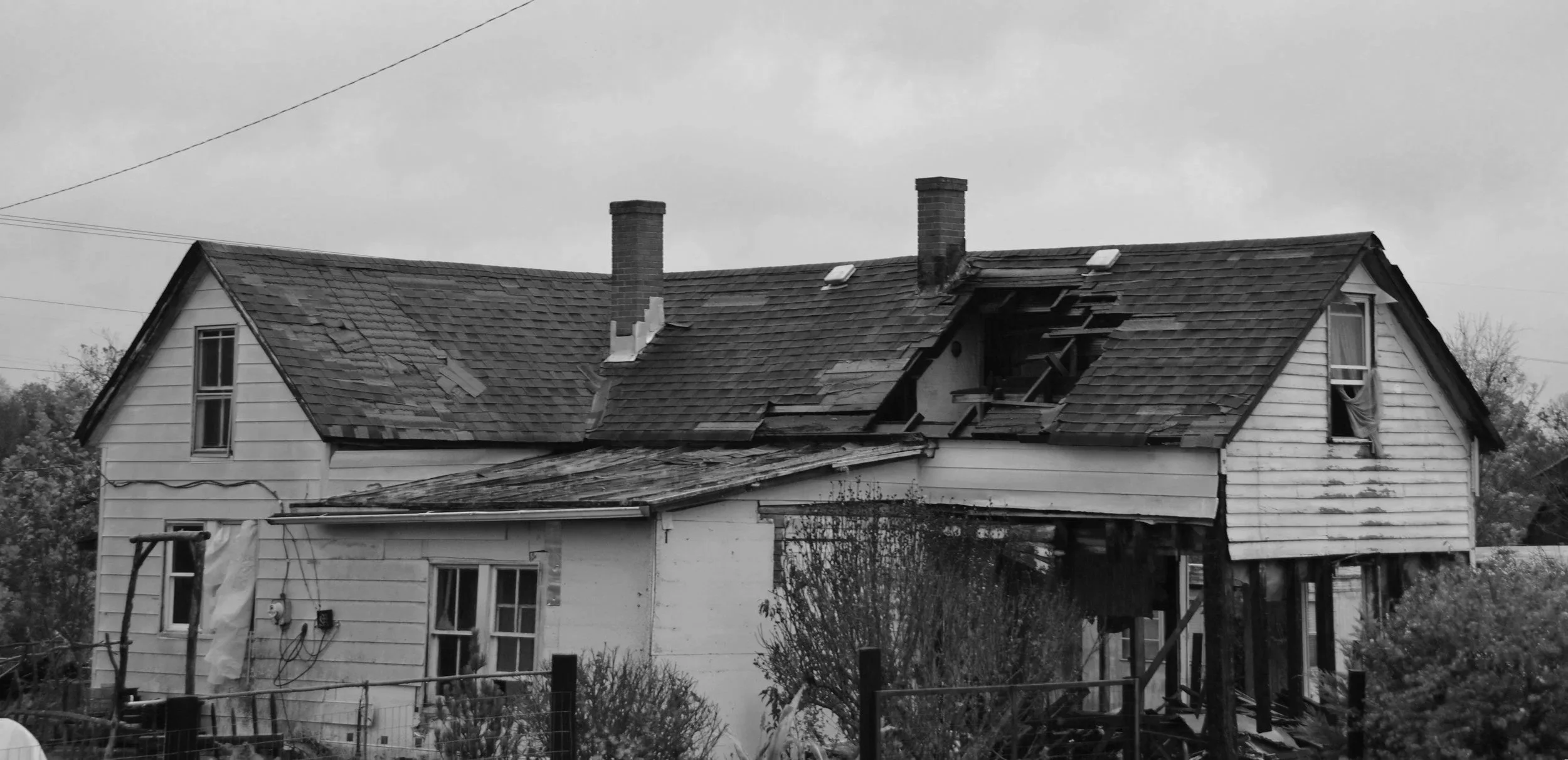A house with extensive roof damage, with missing shingles and a collapsed section on the right side, showing exposed interior and damaged roof structure.