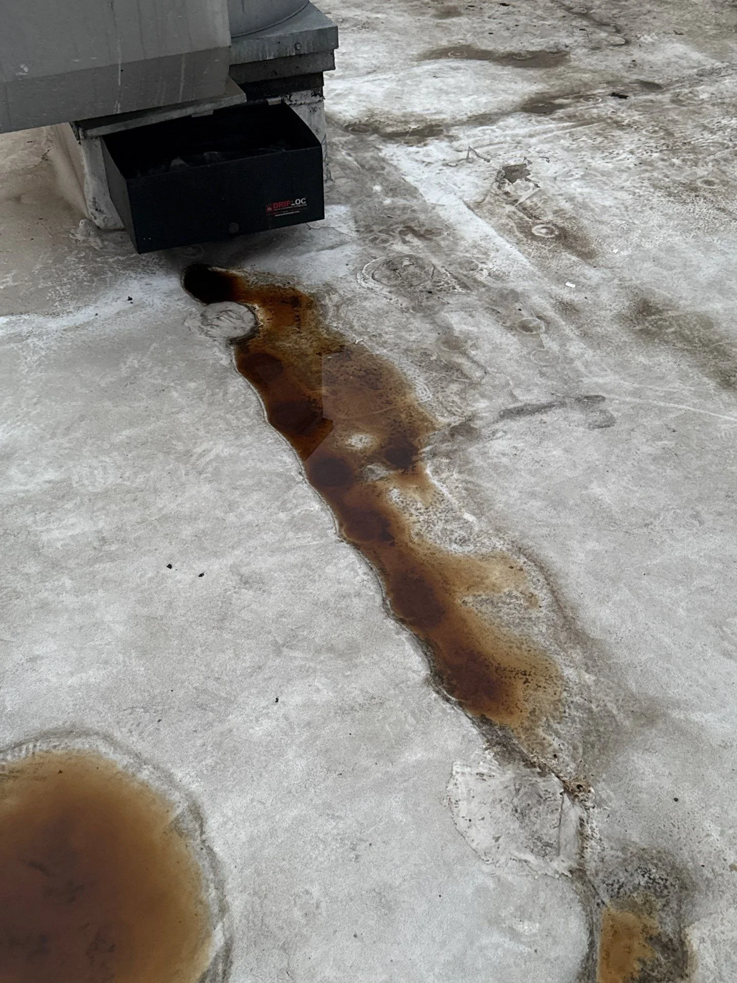 A commercial flat roof with rust-colored liquid spilled from a vent or duct mounted onto the roof. The liquid has pooled and left a stain on the surface.
