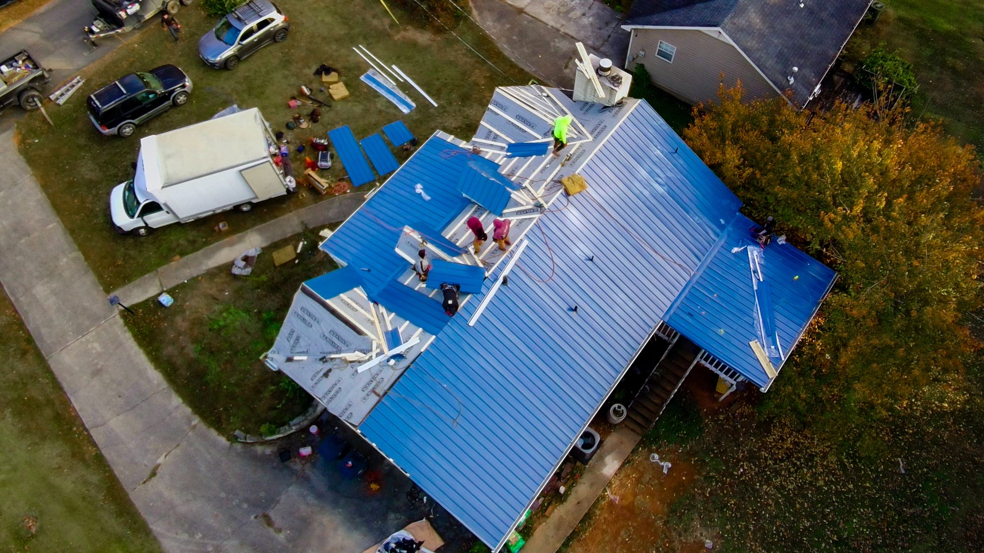 An aerial view of a house under construction with workers installing a new blue metal roof. Tools and materials are scattered around the roof and yard.