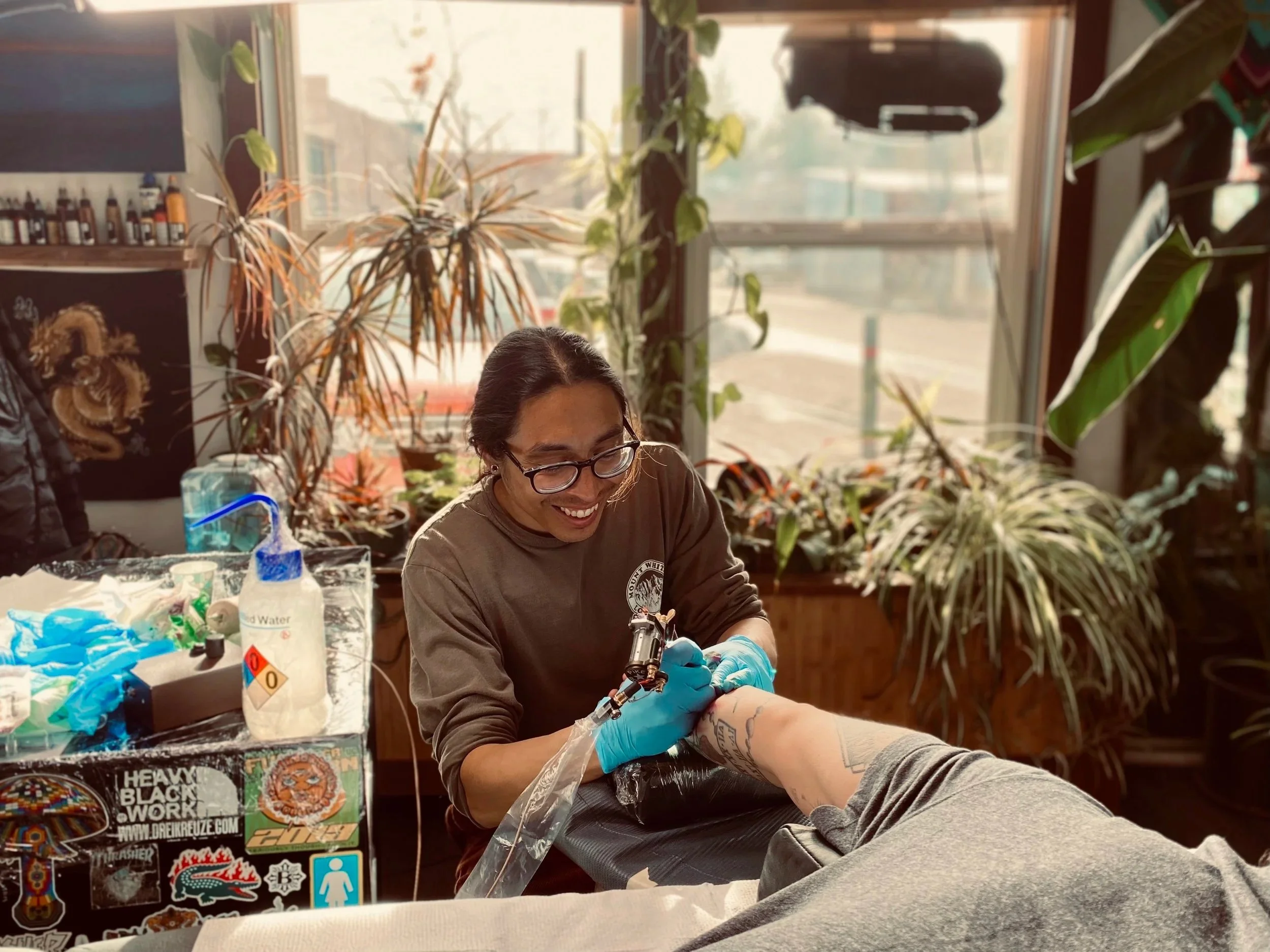 A man with glasses and gloves tattooing a person's arm in a tattoo studio, with plants and supplies visible around, sunlight coming through large windows in the background.
