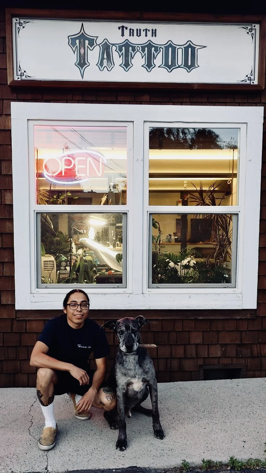 A person with glasses and tattoos crouching beside a large black and gray dog outside a tattoo shop. The shop has a sign that reads 'Truth Tattoo' and a neon sign in the window that says 'Open'.