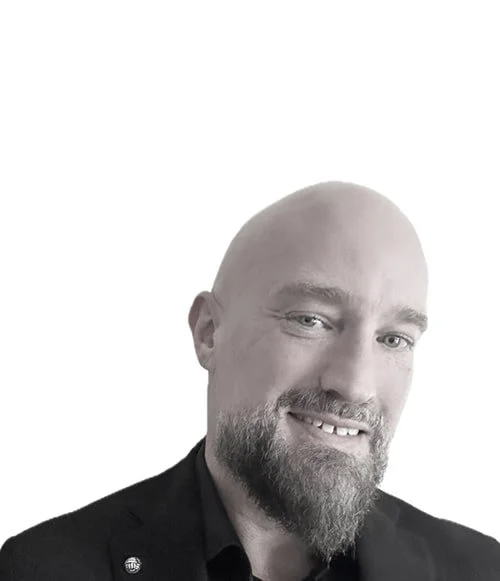 A man with a shaved head and beard smiling, wearing a black blazer against a white background.