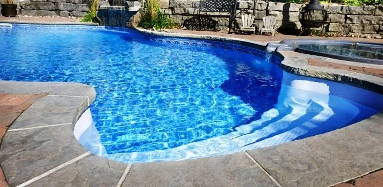 Swimming pool with clear blue water, surrounded by stone and brick patio, with outdoor furniture and a water fountain in the background.