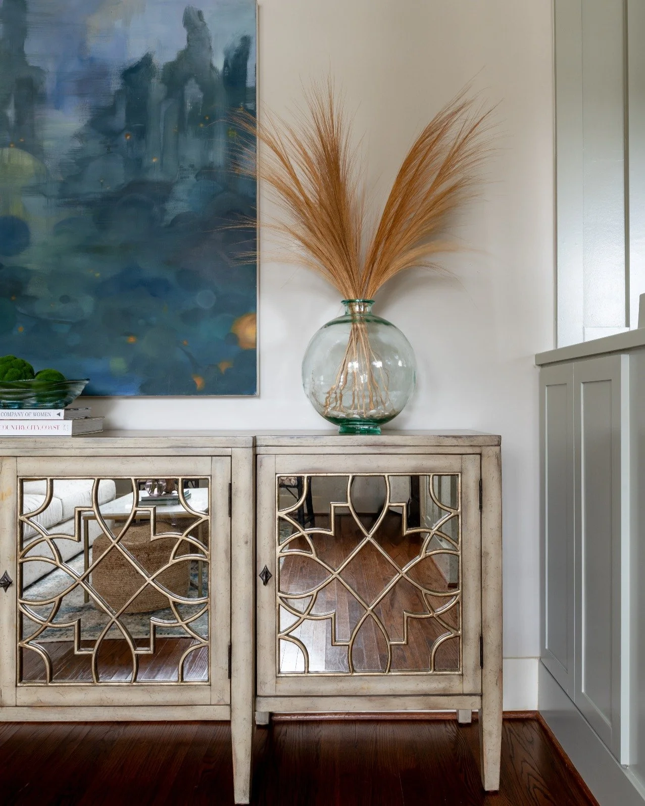 A quiet moment of reflection 
This vignette combines a mirrored console with intricate latticework, a handblown glass vase filled with natural wheat stems, and a moody blue abstract painting. The mix of texture, color, and sheen brings depth to an ot